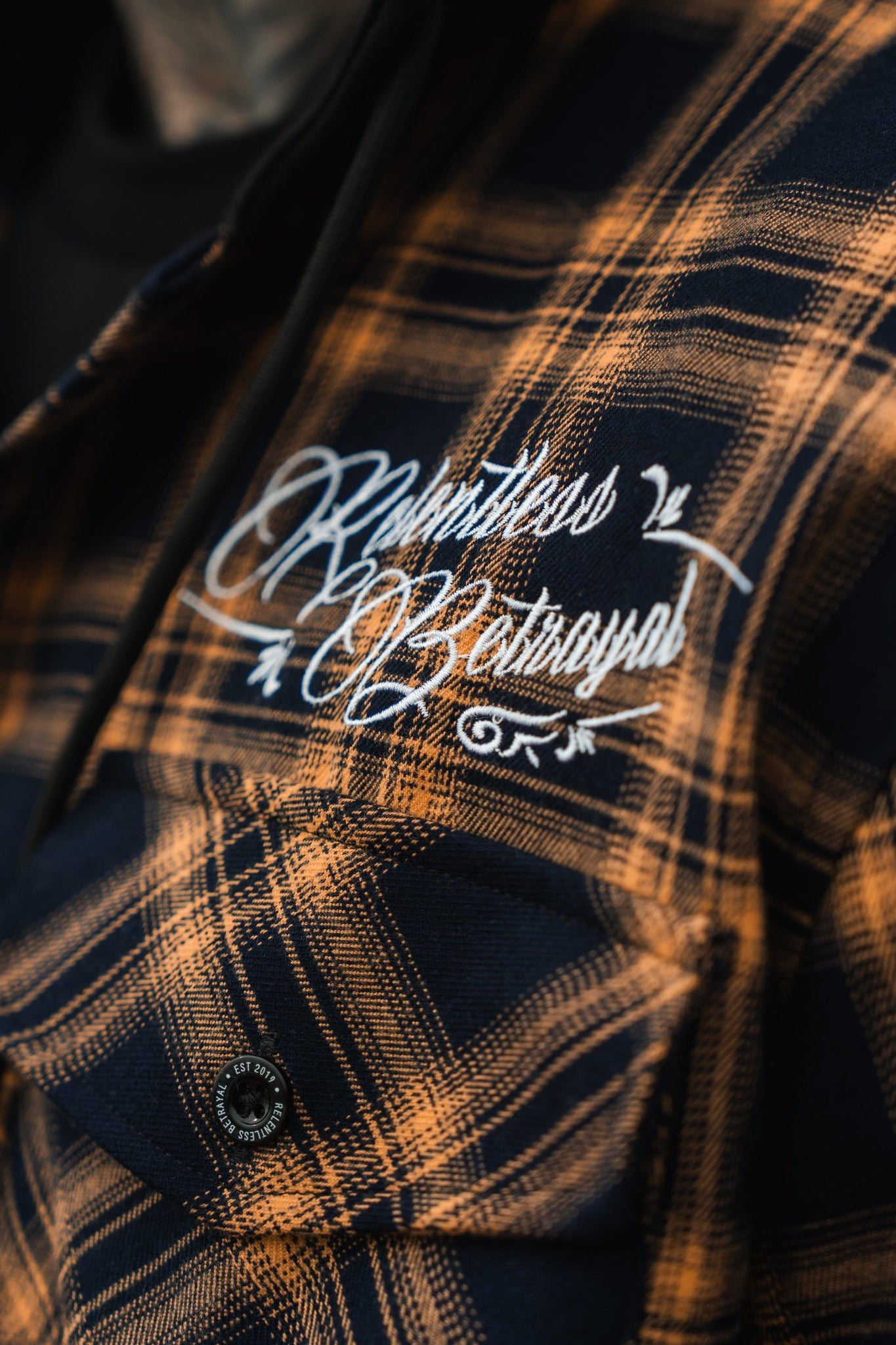 Close-up of someone in a Skilled Sailor Premium Hooded Flannel by Relentless Betrayal, featuring black and yellow plaid. Relentless Betrayal is embroidered in white script on the chest pocket. Made from a cozy cotton/polyester blend, this ultra-soft piece promises comfort and style.