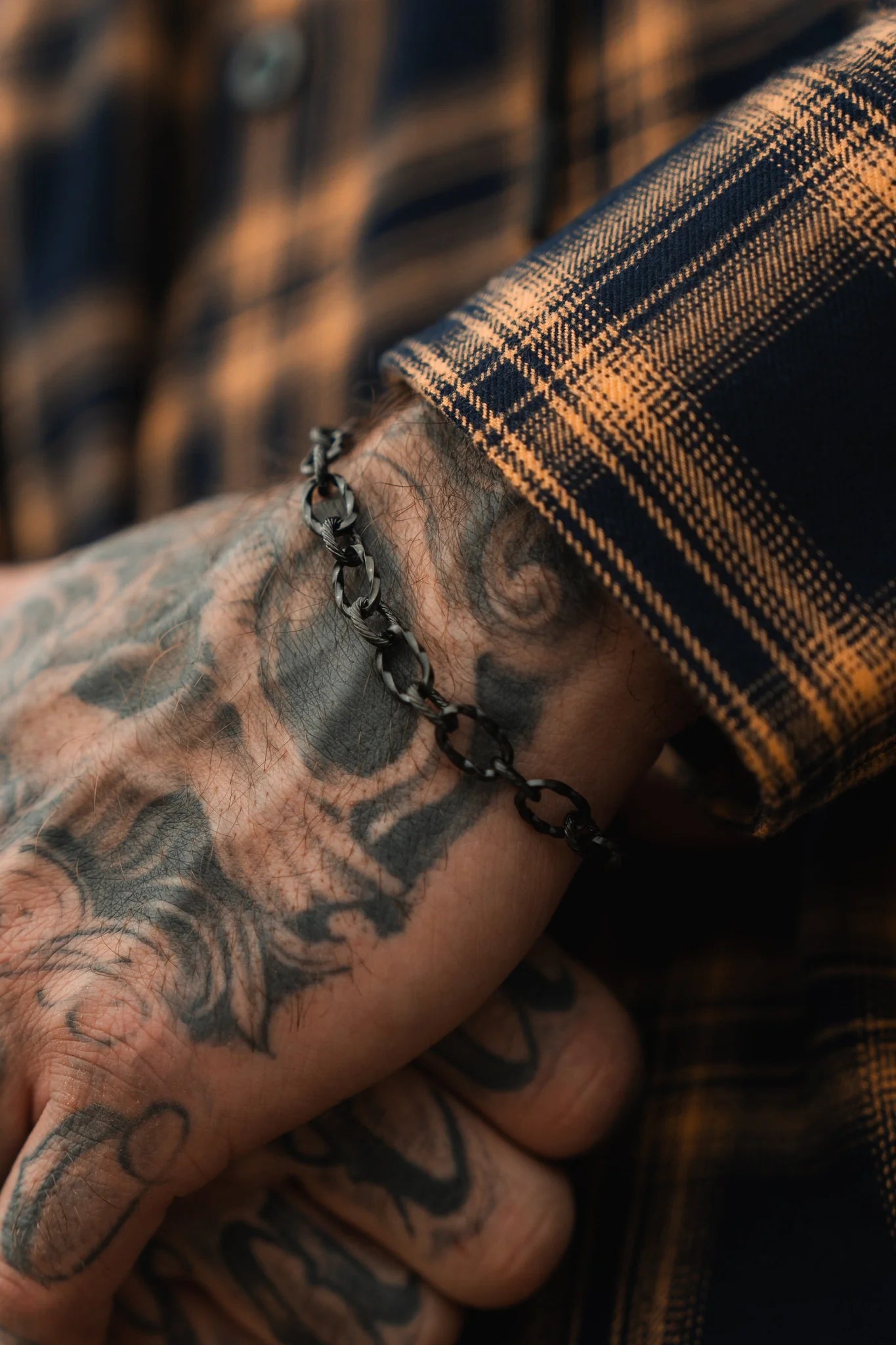 A tattooed hand dons the Mariners Midnight Bracelet by Relentless Betrayal, paired with a plaid orange and black shirt, echoing Dark Seas mystique. The intricate skin designs tell stories akin to maritime legends across the fully adorned hand.