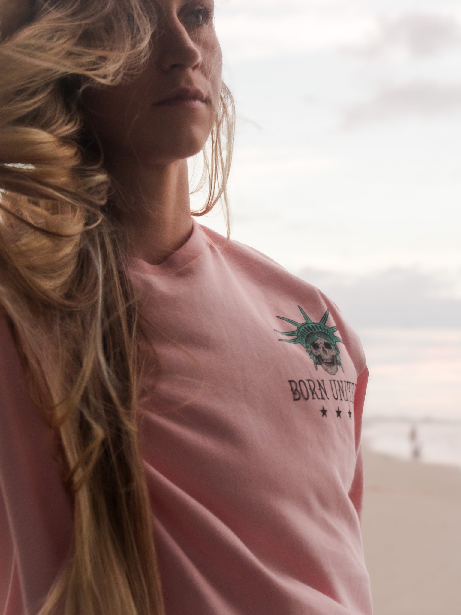 A woman with long blonde hair wears the Born United Private Label Liberty Or Death- Heather Pink tee featuring the Statue of Libertys head and BORN UNIQUE text with stars, standing on a beach at sunset.