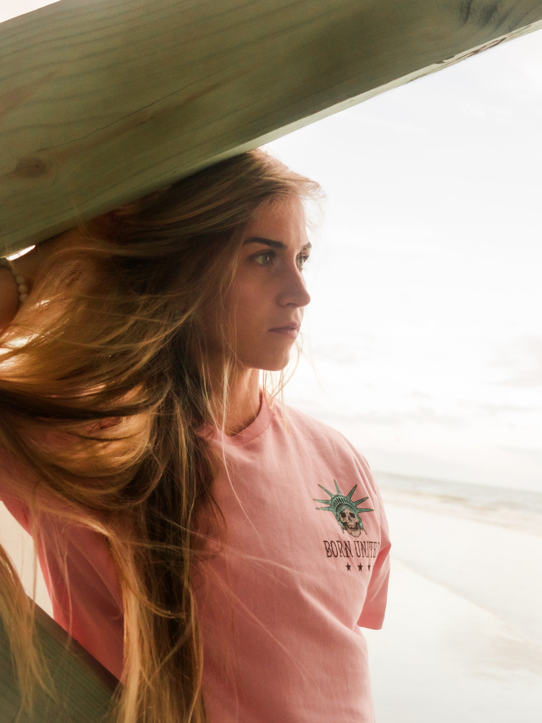 A young woman with long blonde hair stands on the beach under a wooden structure, wearing the Born United Private Label Liberty Or Death- Heather Pink tee. The ocean and a cloudy sky form the scenic background.