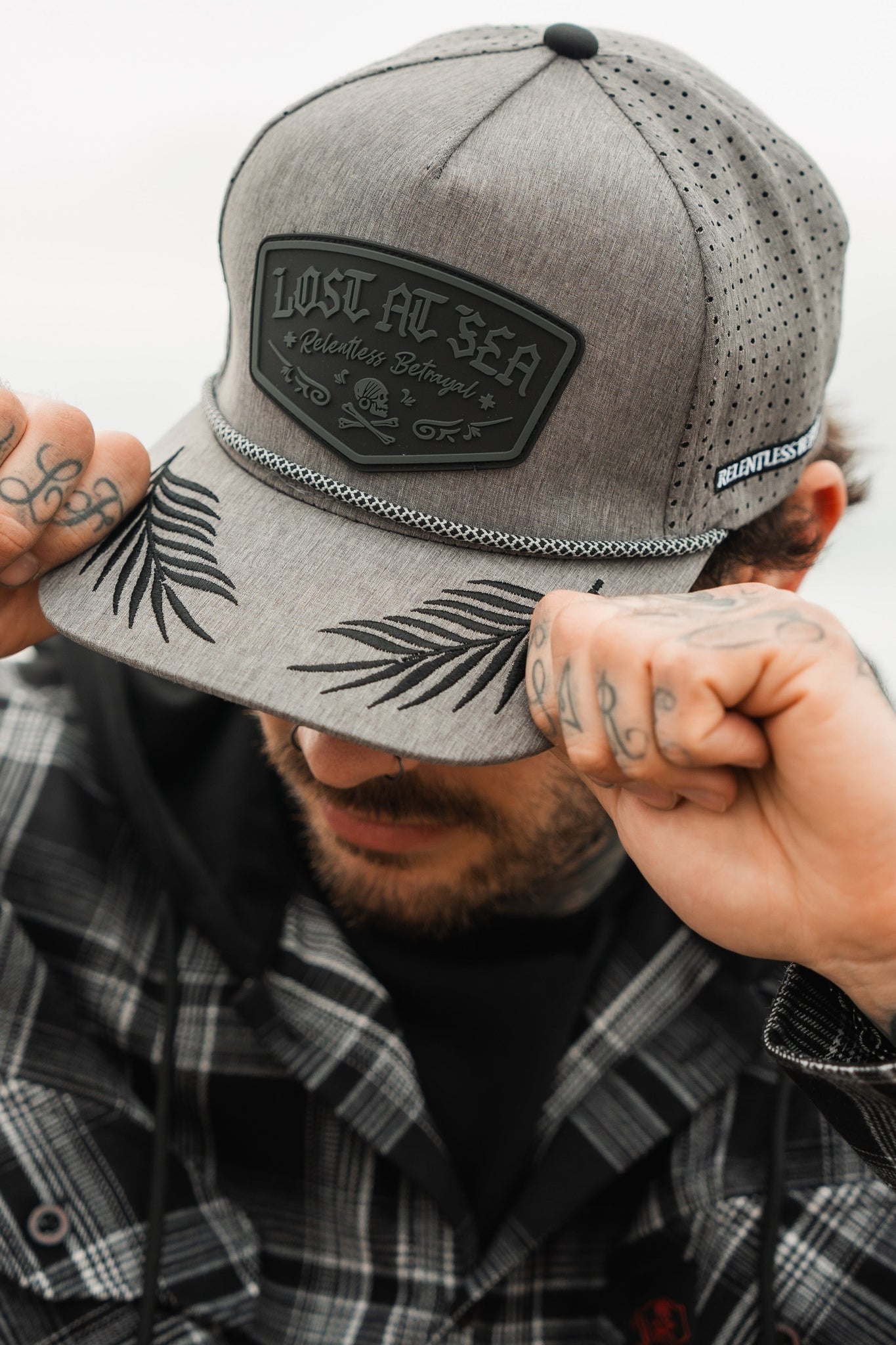 A person in a plaid shirt adjusts the Lost At Sea Gunmetal PVC Snapback by Relentless Betrayal, with LOST AT SEA etched on it. The water-resistant hat features palm leaf designs and perforated sides, as tattooed hands stand out against a softly blurred background.