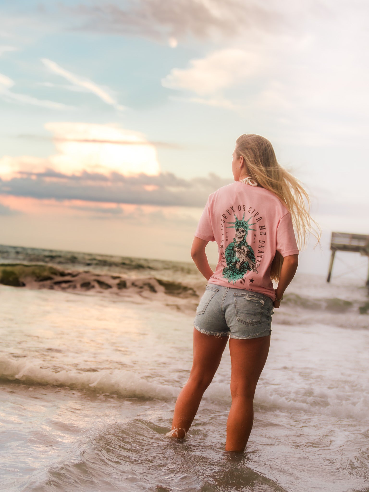 A woman with long blonde hair stands in shallow ocean water at sunset, wearing the Born United Private Label Liberty Or Death- Heather Pink tee and denim shorts, gazing at the horizon with waves around her and a pier behind her.