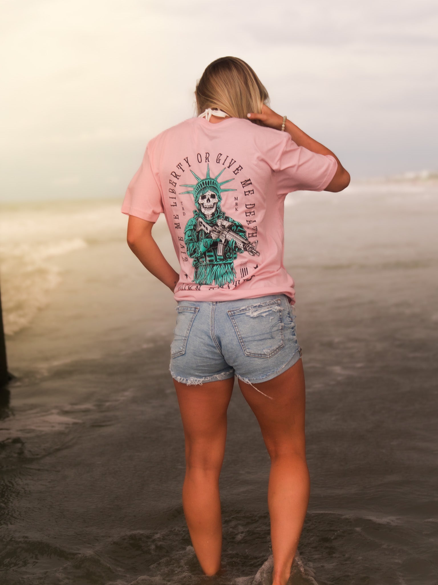 A woman with blonde hair, wearing the Born United Private Label Liberty Or Death- Heather Pink tee, stands in shallow beach water facing away from the camera, paired with denim shorts.
