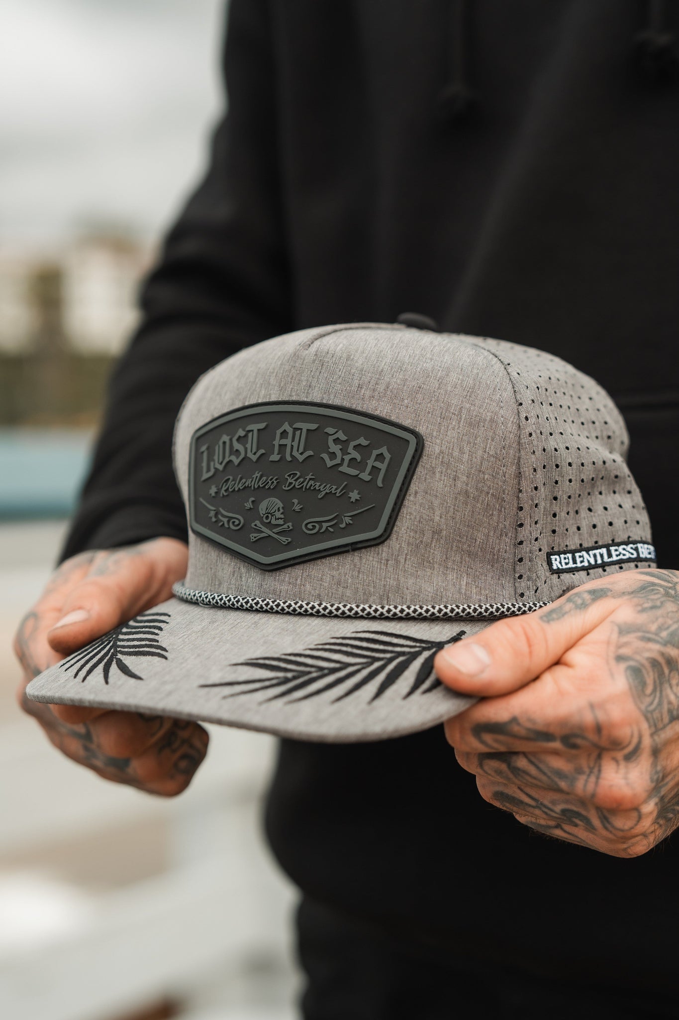 A person with tattooed hands is sporting a custom Lost At Sea Gunmetal PVC Snapback by Relentless Betrayal, showcasing Lost at Sea and Relentless Belong on the front, leaf designs on the brim, and air holes on the side. Theyre also wearing a black hoodie.