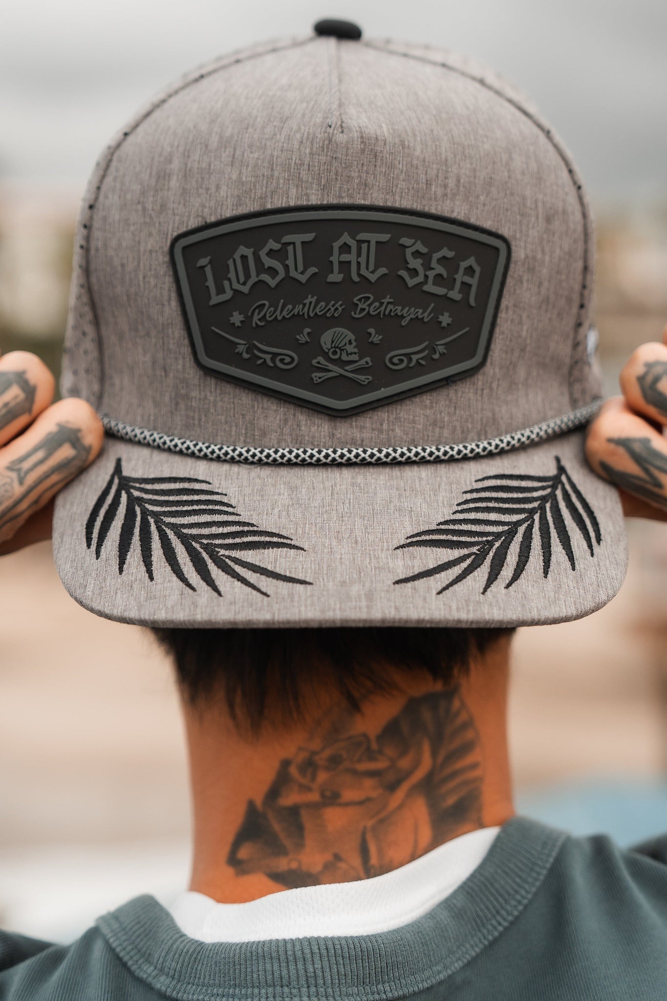 A person with hand tattoos adjusts a grey Lost At Sea Gunmetal PVC Snapback by Relentless Betrayal. This water-resistant cap has embroidered black leaves on the brim and a skull and crossbones patch reading LOST AT SEA Relentless Betrayal.