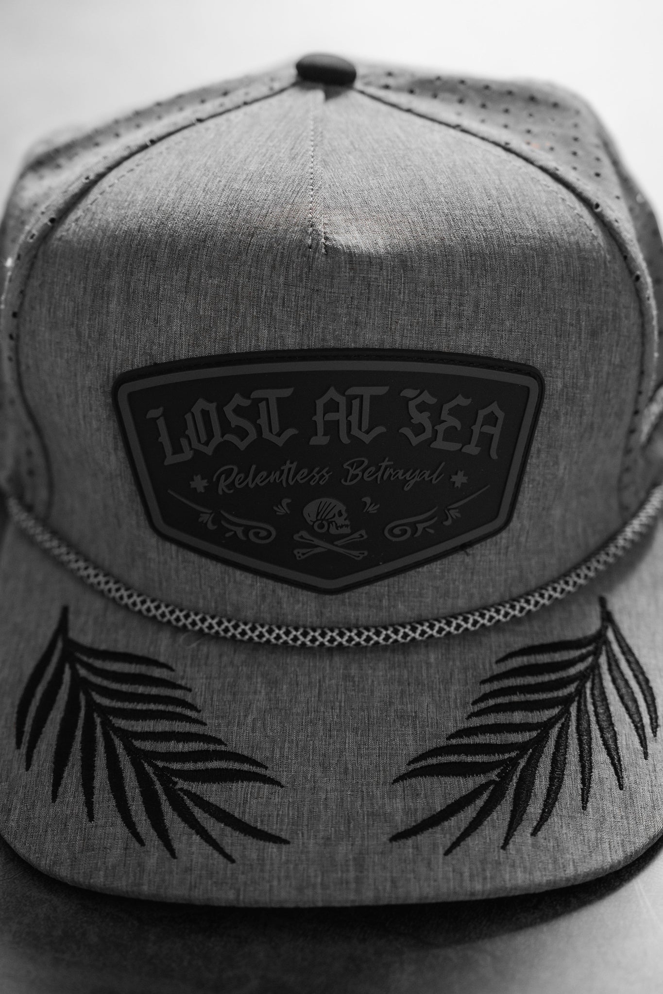 The Lost At Sea Gunmetal PVC Snapback by Relentless Betrayal is a custom grey hat with black embroidered palm leaves on the brim, featuring a LOST AT SEA front patch. Its a water-resistant snapback capturing its unique charm in black-and-white with Relentless Betrayal and an emblem.