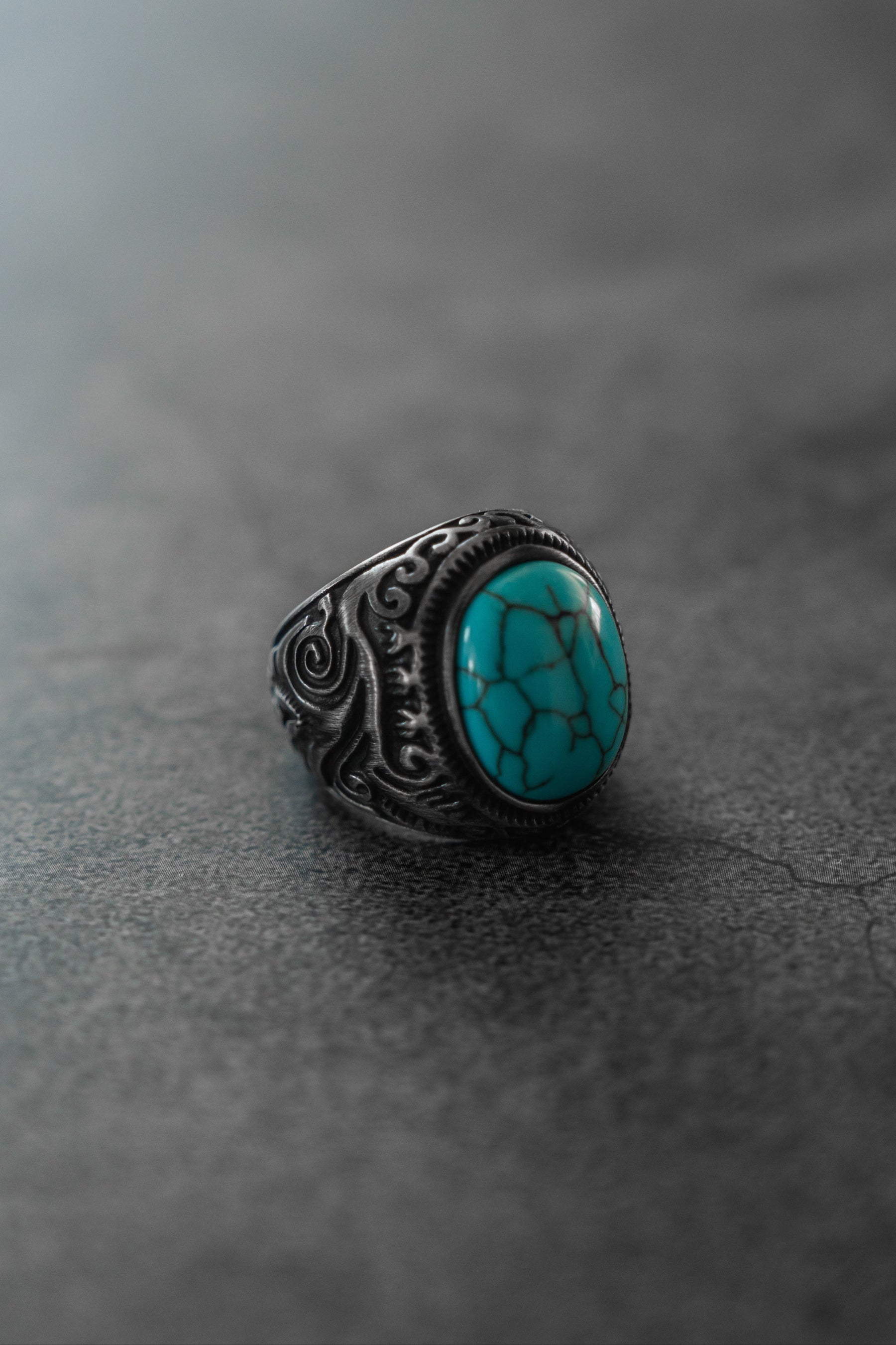 The Into the Storm Ring by Relentless Betrayal showcases a large oval turquoise stone with black veining and ornate silver designs, beautifully displayed on a smooth, gray, stone-like surface.