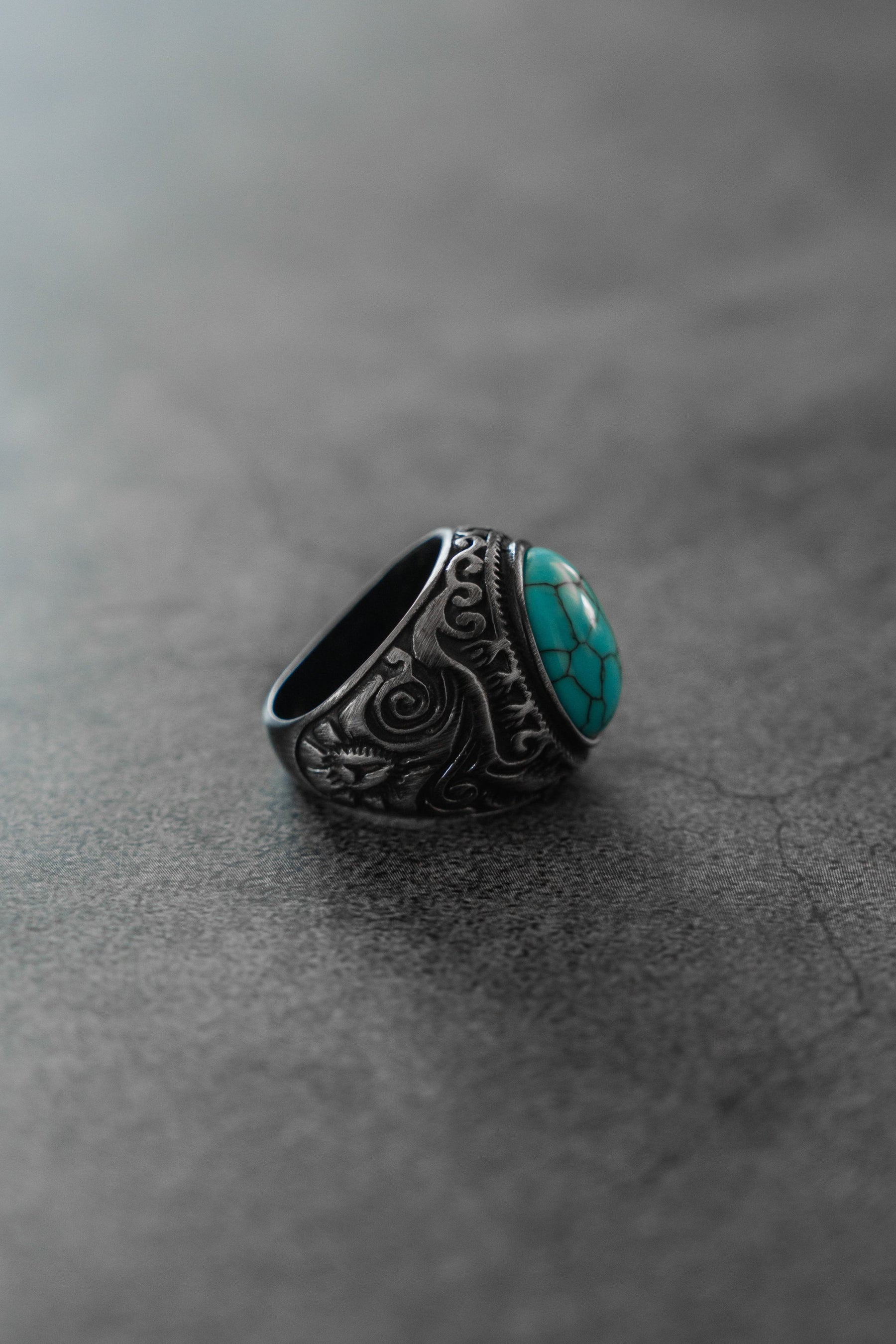 The Into the Storm Ring by Relentless Betrayal features intricate engravings on handcrafted stainless steel and a bold turquoise stone, displayed on a smooth gray surface.