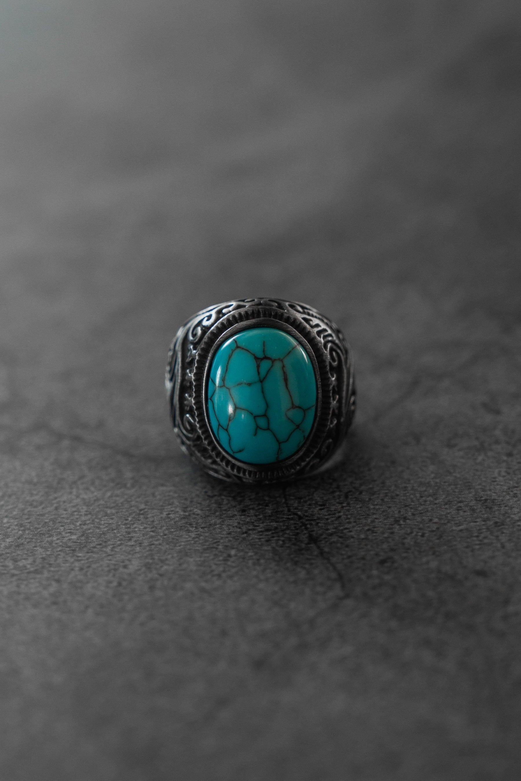 The Relentless Betrayal Into the Storm Ring features silver with ornate engravings and a large oval turquoise stone with black veining, showcased on a dark, textured surface.