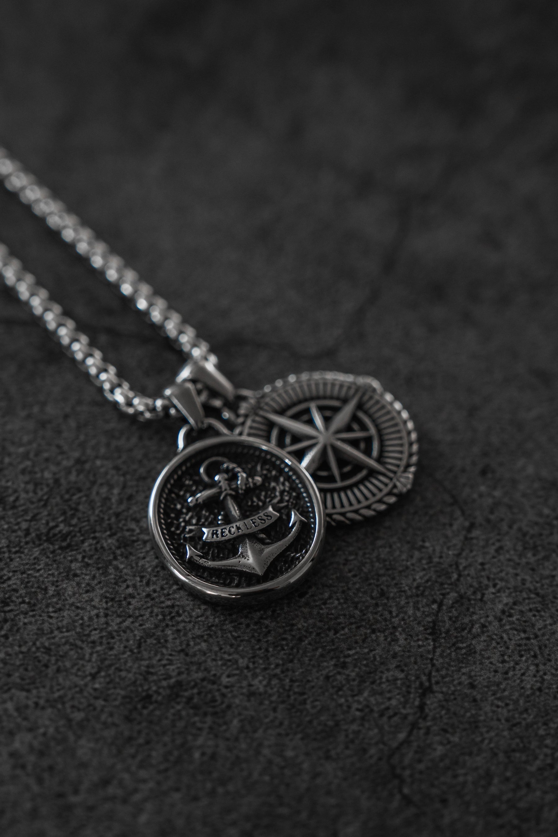 Two silver stainless steel necklaces from Relentless Betrayal: the Reckless Soul Necklace with an anchor engraved RECKON, and a compass pendant. Both handcrafted pieces rest on a dark textured surface.