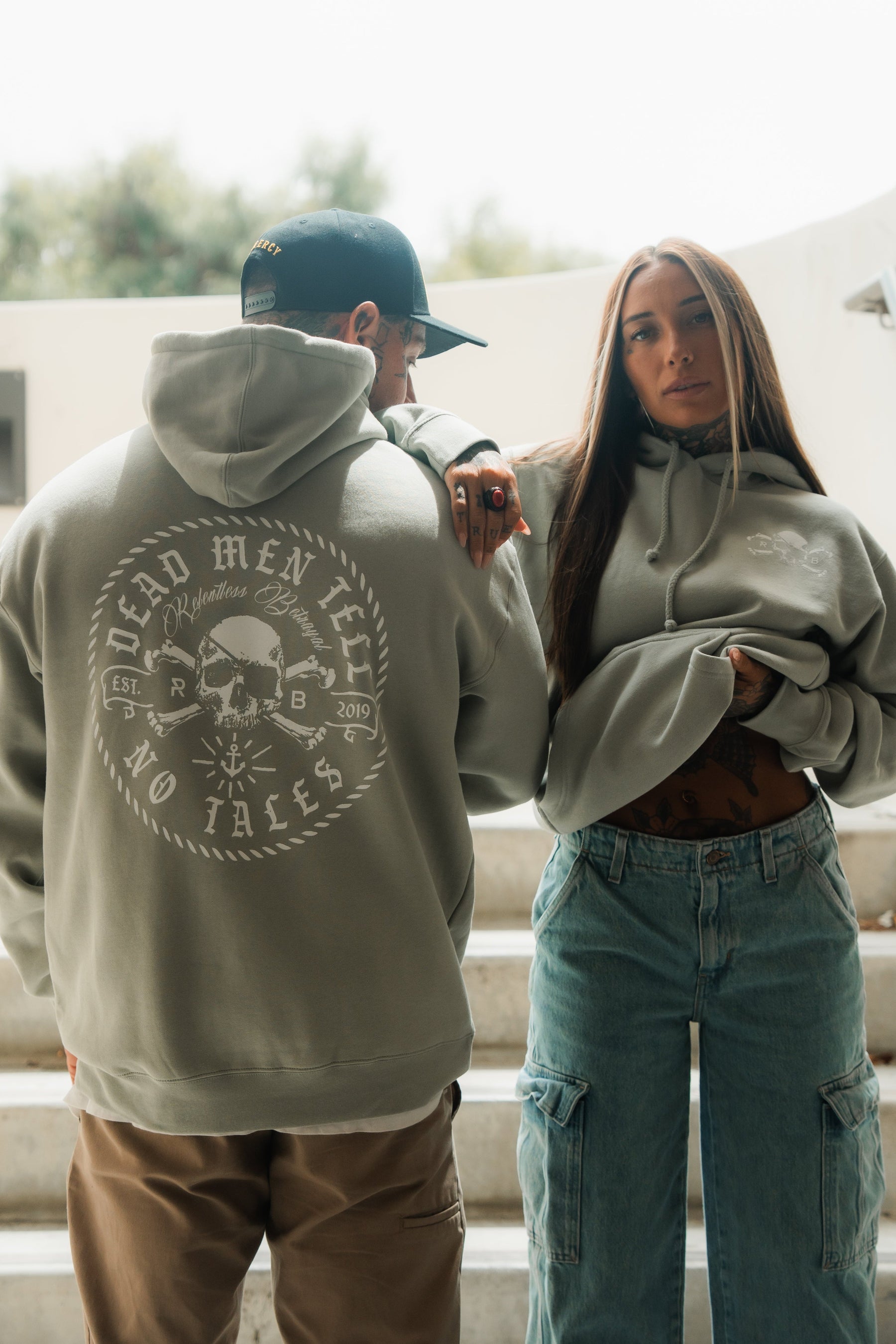 Two people wear Relentless Betrayals Tell No Tales Premium Sage Hoodie; the man shows off a bold t-shirt design, while the woman lifts her hoodie to reveal loose blue jeans. Both sport comfortable cotton-polyester blend styles.