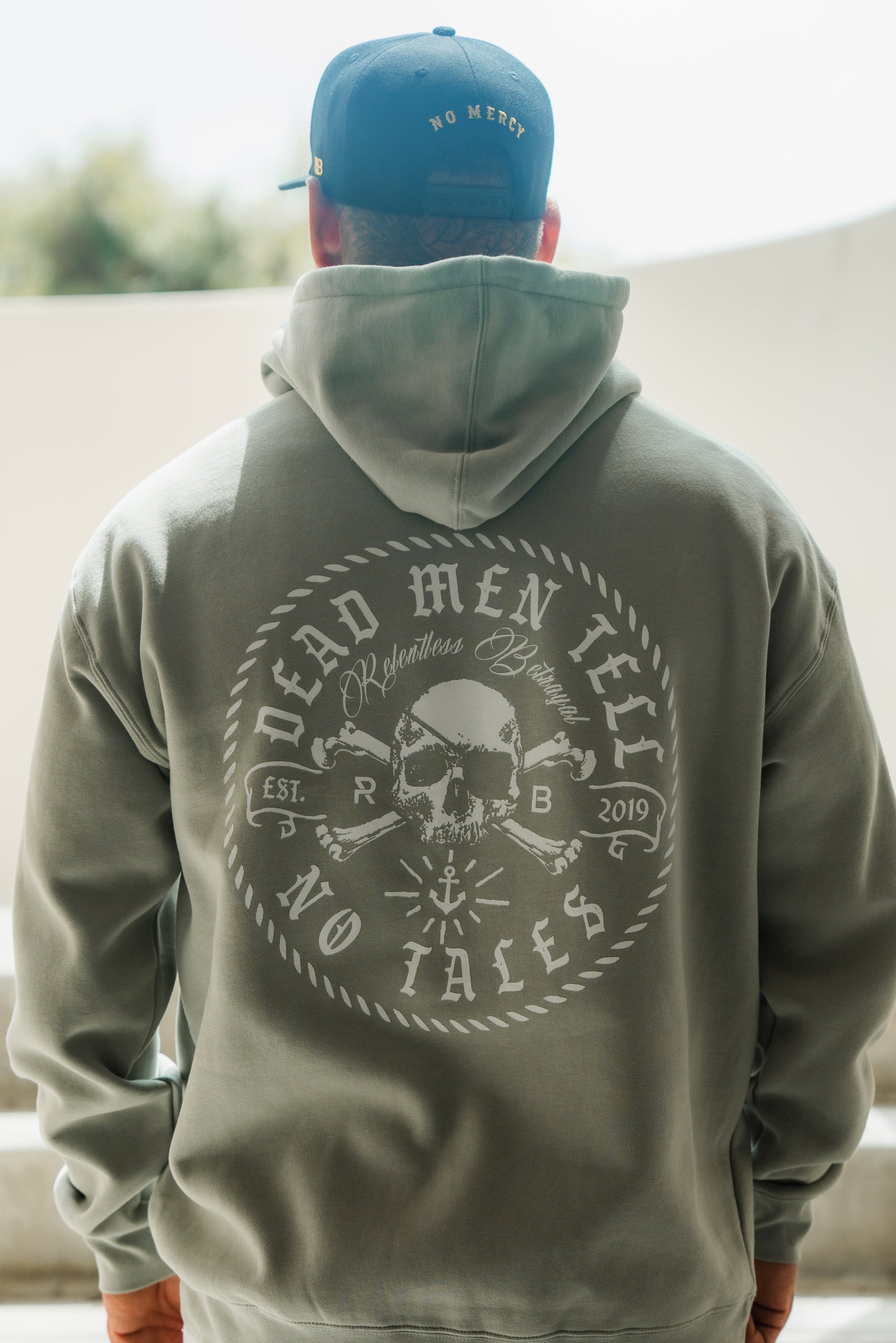 Someone stands outdoors wearing the Relentless Betrayal Tell No Tales Premium Sage Hoodie with a bold white skull and crossbones graphic, paired with a navy cap featuring NO MERCY embroidered on the back.