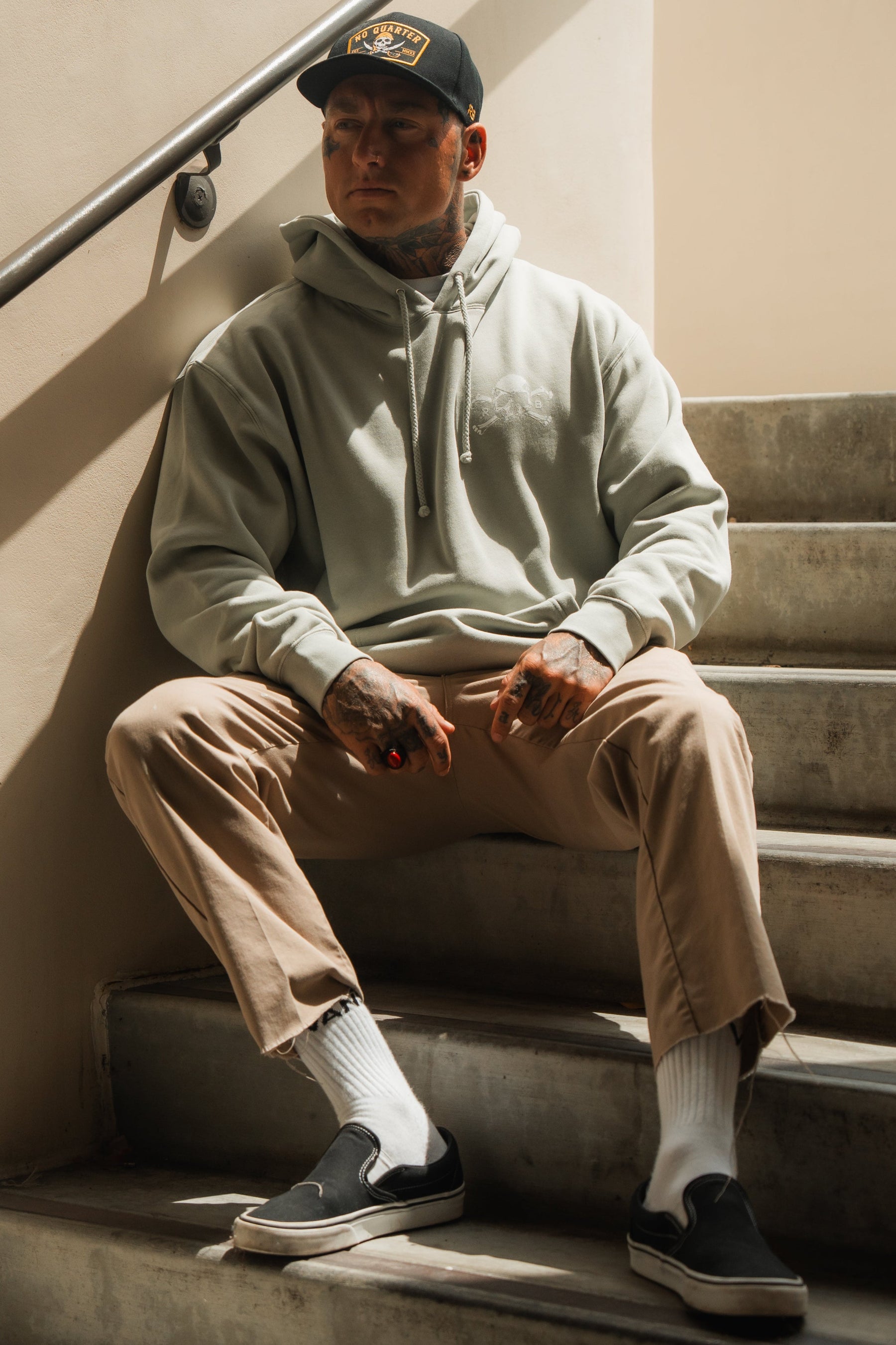A man wearing the Relentless Betrayal Tell No Tales Premium Sage Hoodie with tan pants and a black cap sits on concrete stairs. His tattoos show as he looks away, pairing black slip-ons and white socks for a laid-back, stylish vibe.