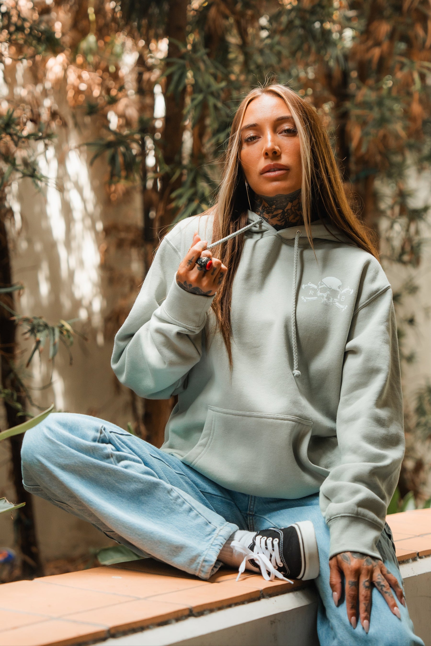 A woman with long hair and tattoos sits cross-legged outdoors on a ledge, wearing the Relentless Betrayal Tell No Tales Premium Sage Hoodie and blue jeans. She holds a hoodie string and looks at the camera, with trees and plants in the background.
