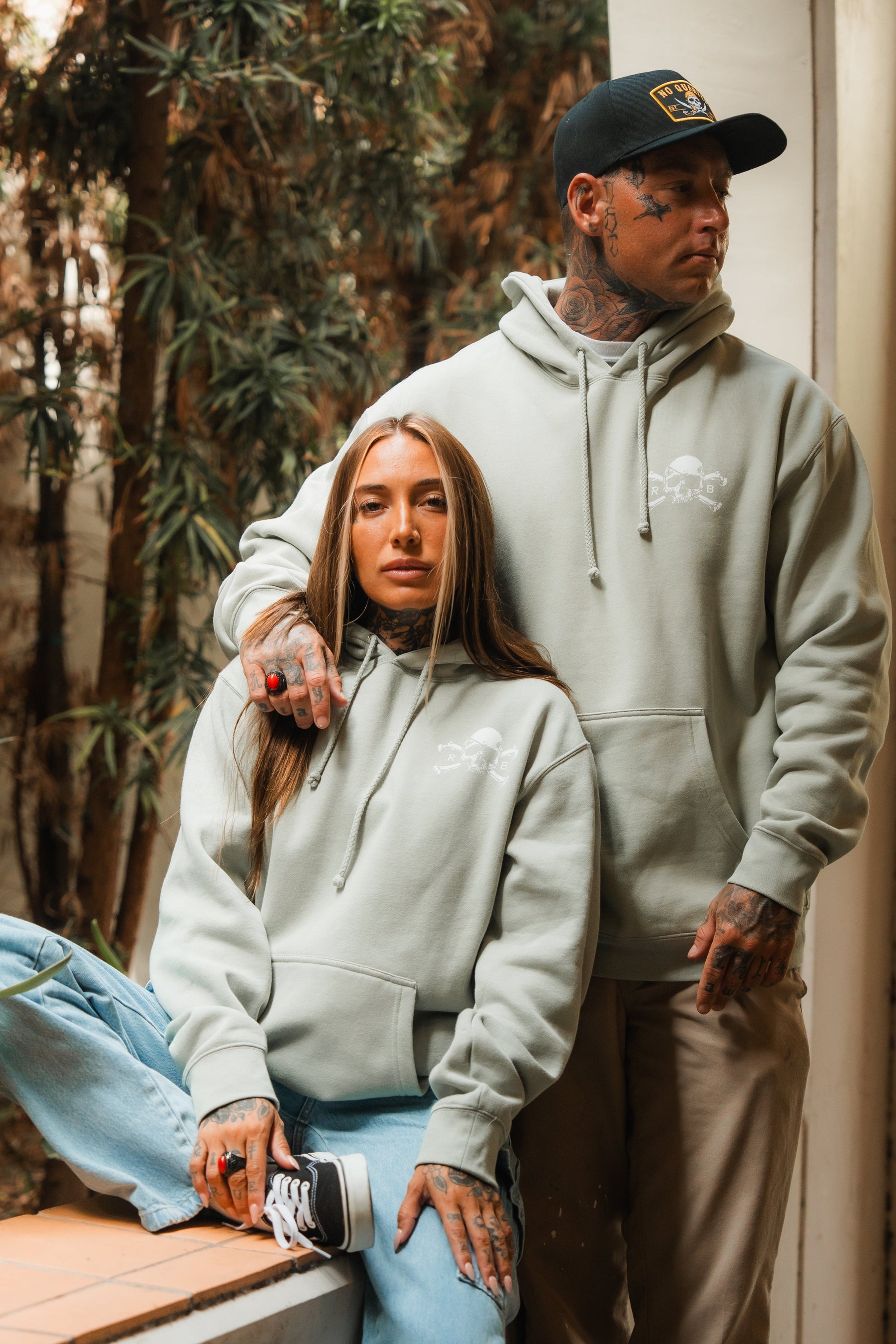 A tattooed woman and man wear matching Relentless Betrayal Tell No Tales Premium Sage Hoodies. She sits on a ledge with one knee up; he stands beside her in a black cap. Both look relaxed and confident.