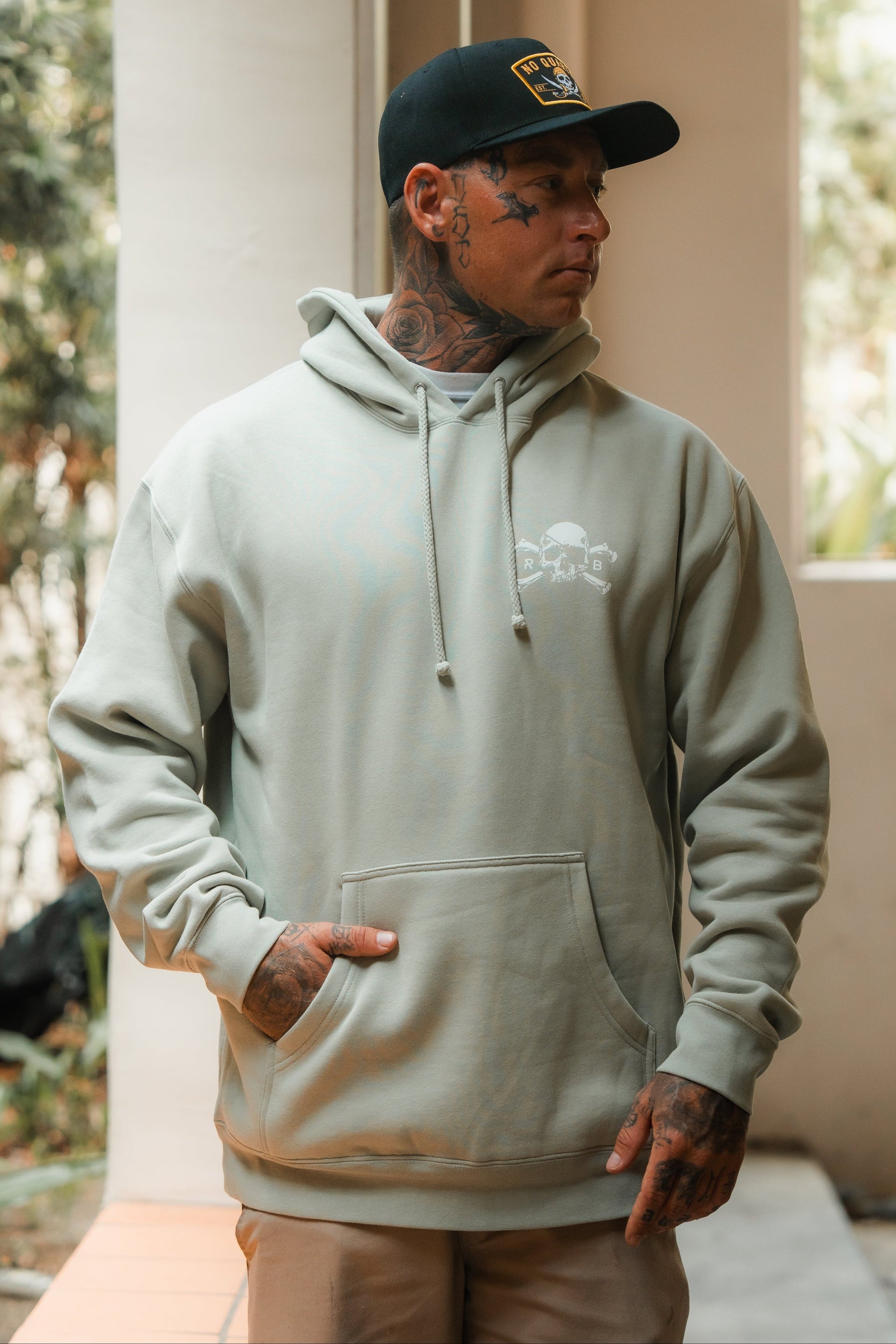 A tattooed man wearing the Relentless Betrayal Tell No Tales Premium Sage Hoodie and a black cap stands indoors, looking to his right with his left hand in the hoodie pocket.