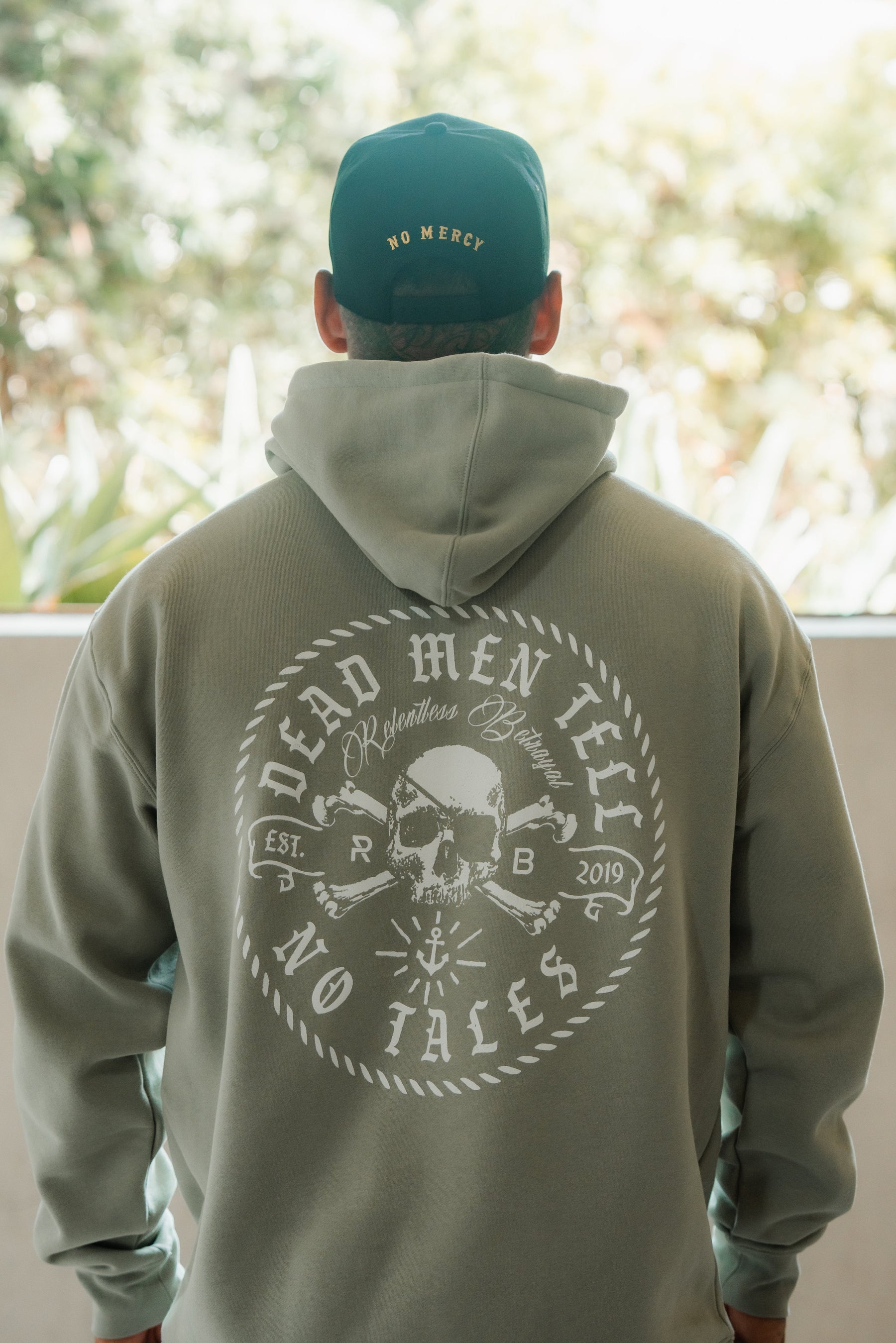 A person wearing a Relentless Betrayal Tell No Tales Premium Sage Hoodie, standing outdoors.
