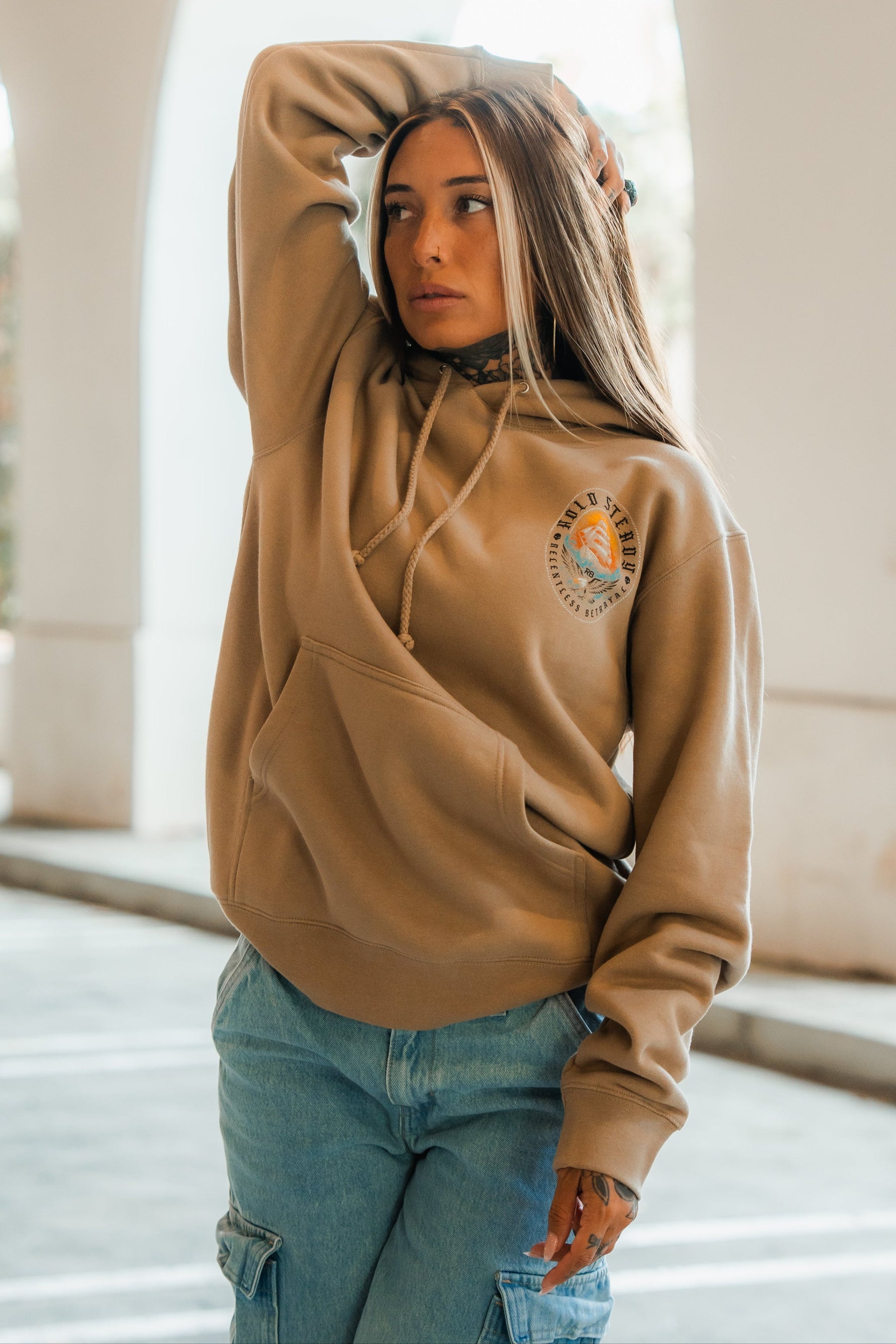 A woman with long hair wears a tan Relentless Betrayal Hold Steady Premium Hoodie and blue cargo jeans, standing outdoors with one arm raised, looking off to the side in the sunlight.
