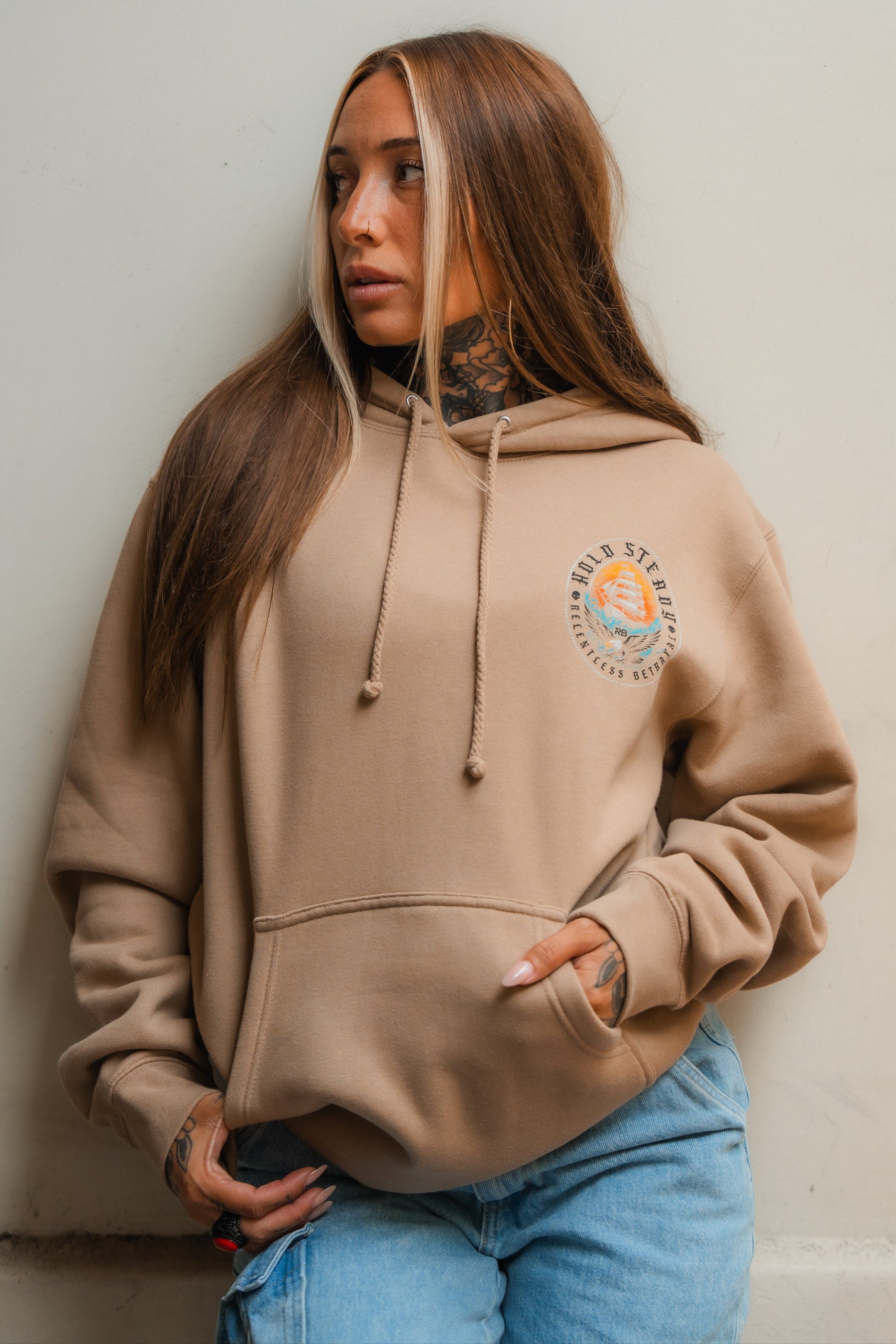 A woman with long brown hair and tattoos wears a Relentless Betrayal Hold Steady Premium Hoodie with a printed emblem and light blue jeans, standing against a plain light-colored wall, looking to her left.