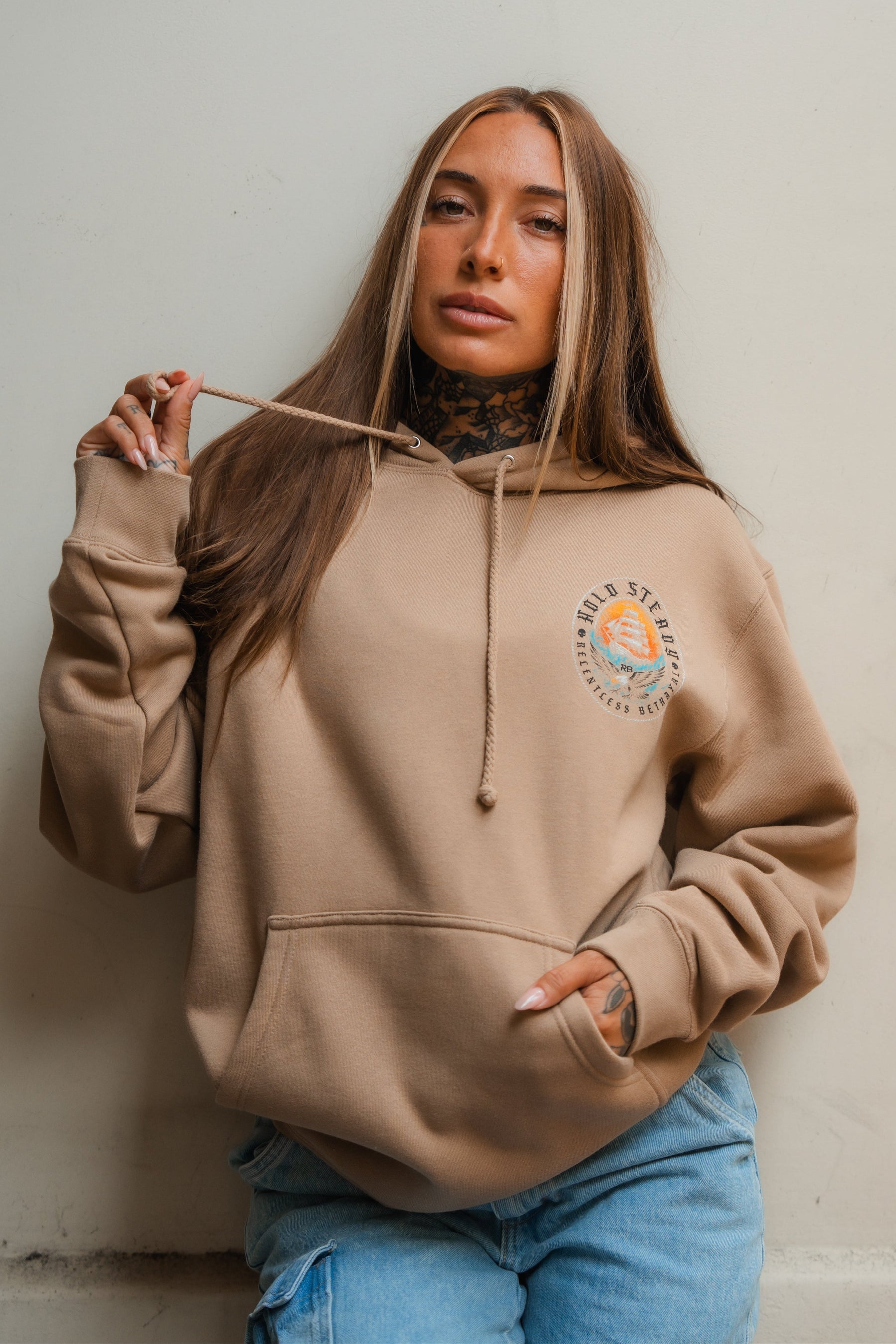 A woman with long, straight hair wears the Relentless Betrayal Hold Steady Premium Hoodie in beige and light blue jeans. She stands against a plain background, holding one drawstring with a neutral expression and one hand in her pocket.