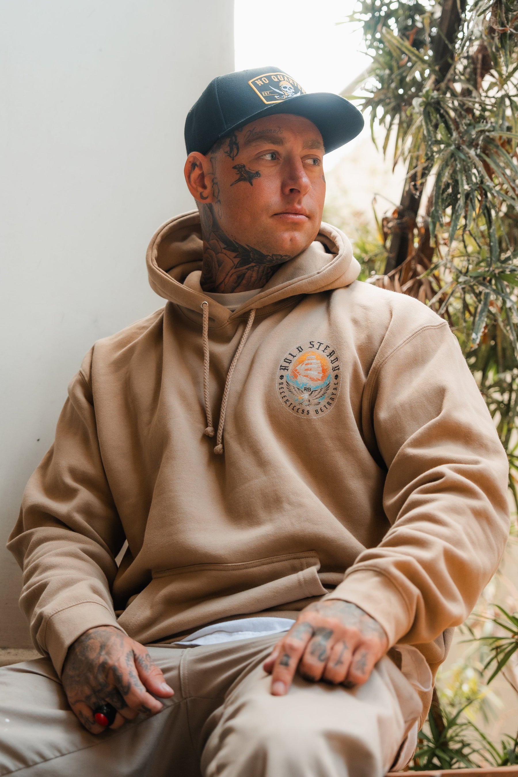 A tattooed man sits indoors by a plant, wearing the Hold Steady Premium Hoodie from Relentless Betrayal in beige and a black cap, looking to the side with a relaxed expression.