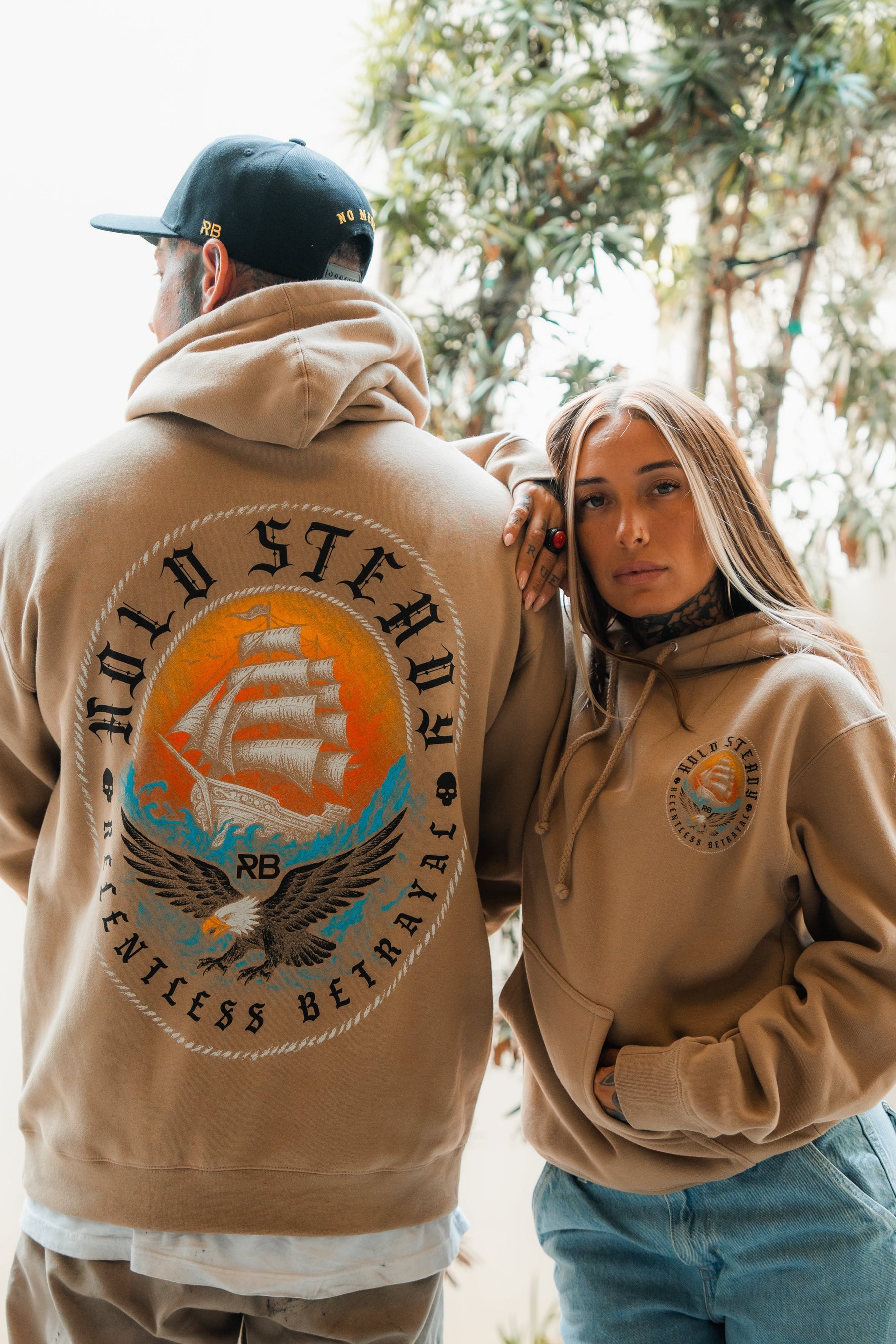 Two people wear Relentless Betrayal Hold Steady Premium Hoodies outdoors. The woman faces forward, while the man shows off the ship graphic and Relentless Betrayal text on the back among leafy trees.