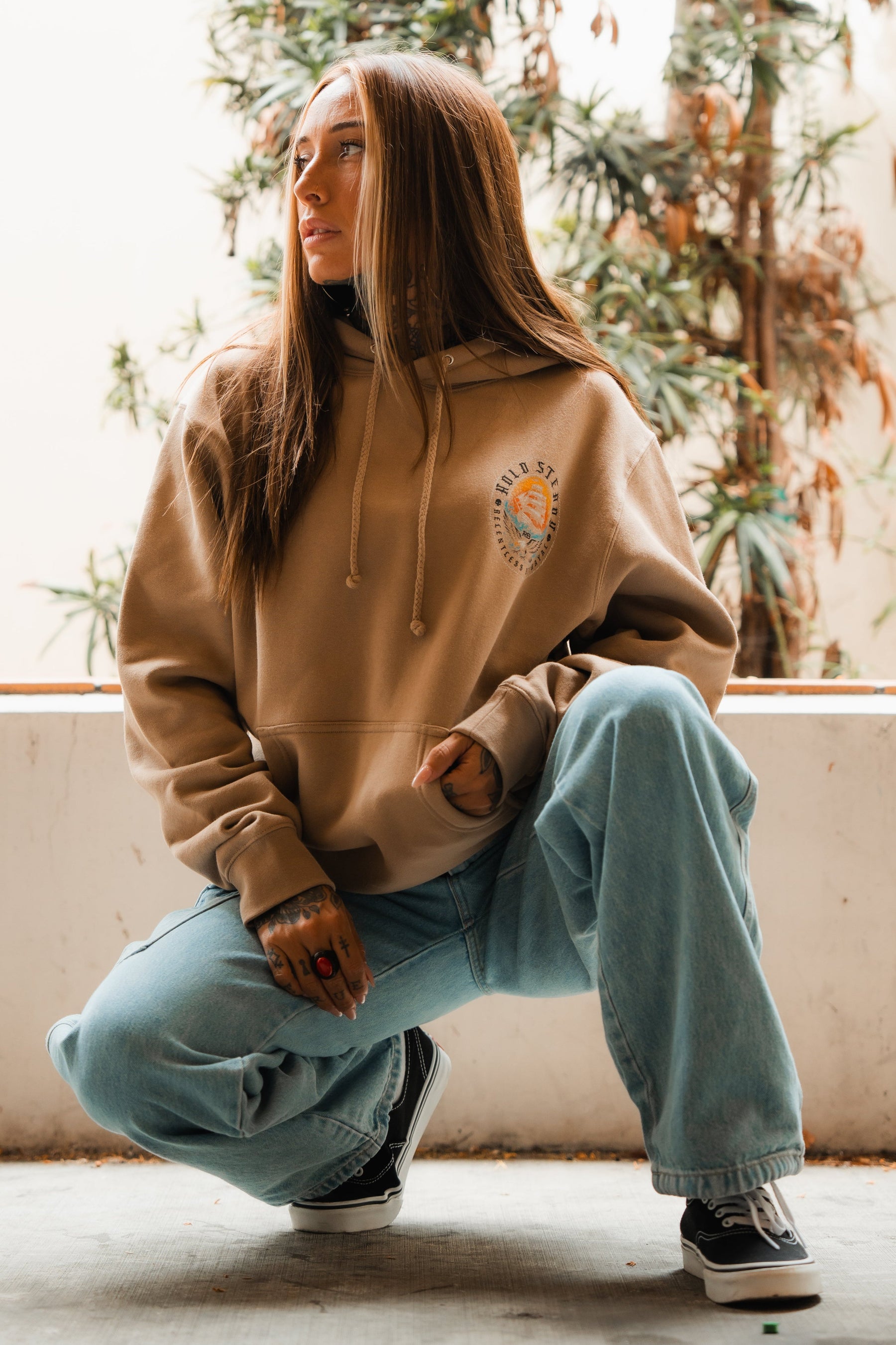 A woman with long brown hair, wearing the Relentless Betrayal Hold Steady Premium Hoodie, light blue baggy jeans, and black-and-white sneakers, kneels outside before leafy plants, gazing to the side with a relaxed expression.