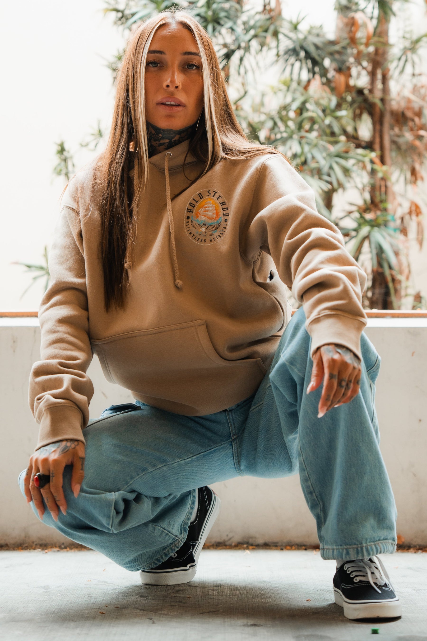 A person with long brown hair and tattoos squats indoors wearing the Relentless Betrayal Hold Steady Premium Hoodie in beige, light blue jeans, and black-and-white sneakers. Green plants can be seen in the background.