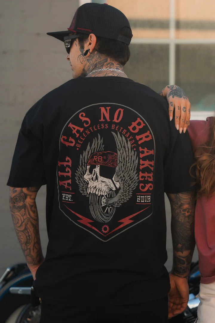 A person wears a black All Gas No Brakes Premium T-Shirt by Relentless Betrayal, featuring a skull and wings design. With tattoos visible and a cap on, they embody youthful edginess as they stand back to the camera.