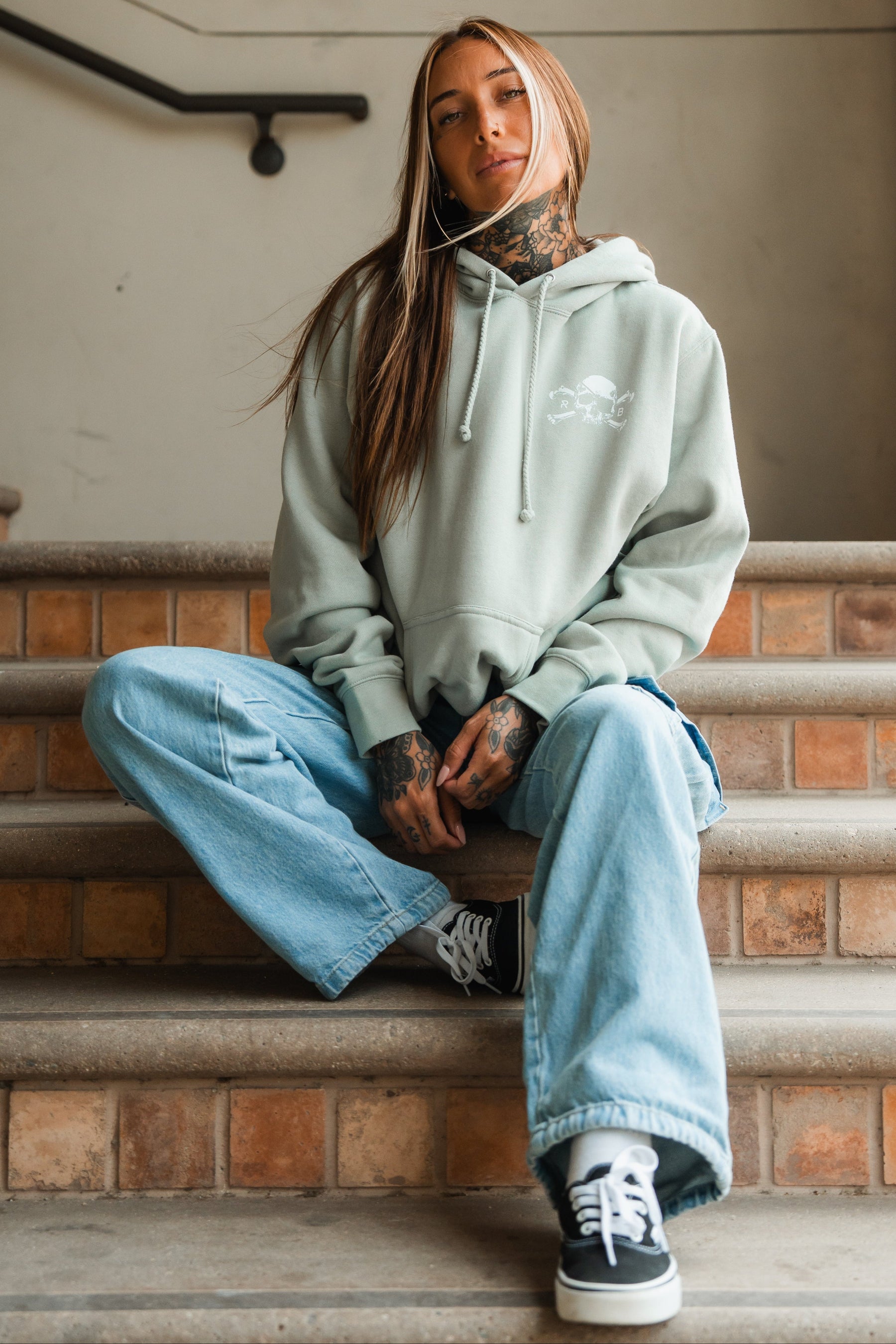 A woman with long hair and tattoos sits on stairs, confidently wearing the Relentless Betrayal Tell No Tales Premium Sage Hoodie, baggy blue jeans, and black sneakers, with one leg raised and hands resting on her knee.