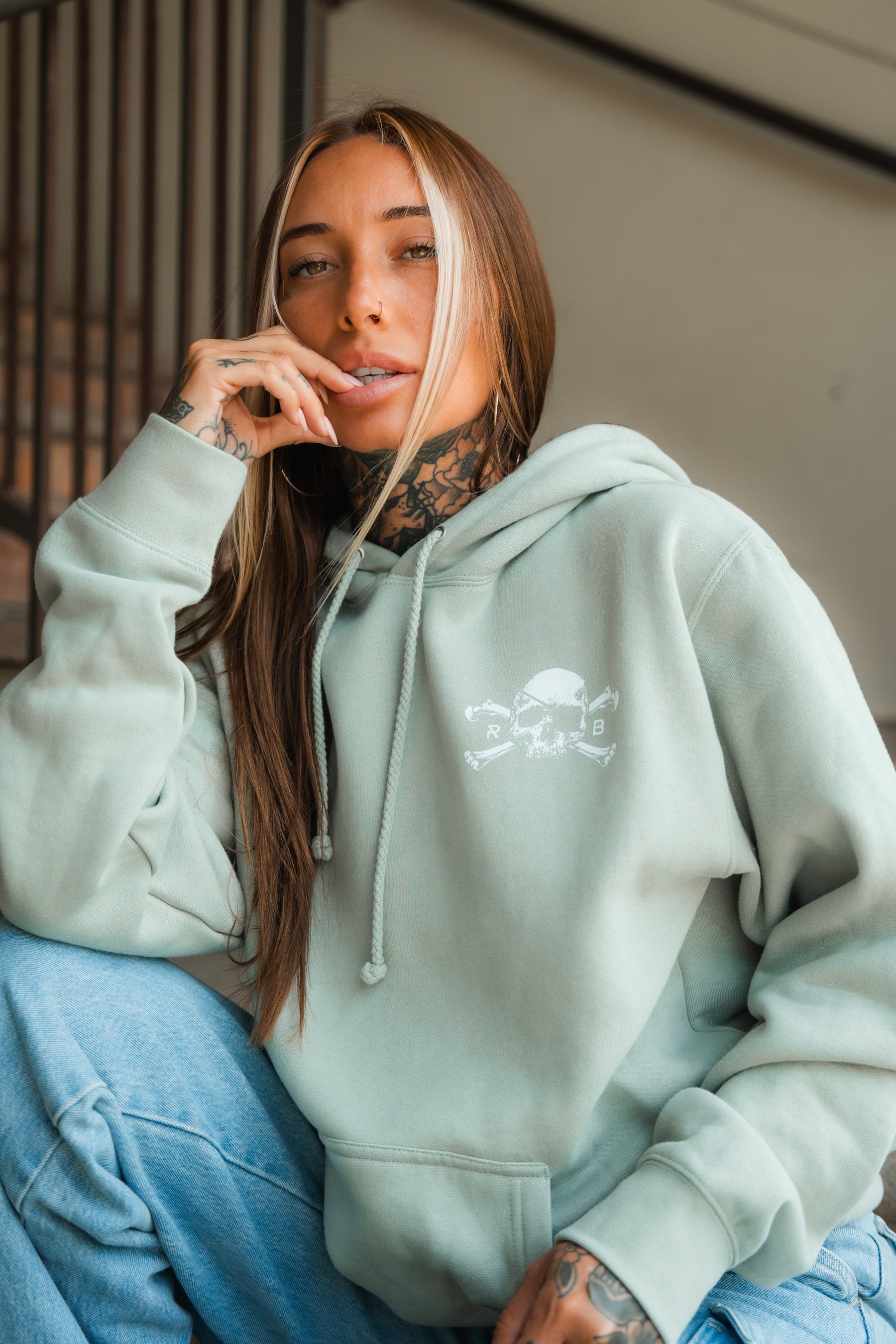 A woman with long brown hair and tattoos poses indoors, wearing the Relentless Betrayal Tell No Tales Premium Sage Hoodie, featuring a white skull graphic, and light blue jeans. She sits casually with her hand near her mouth.