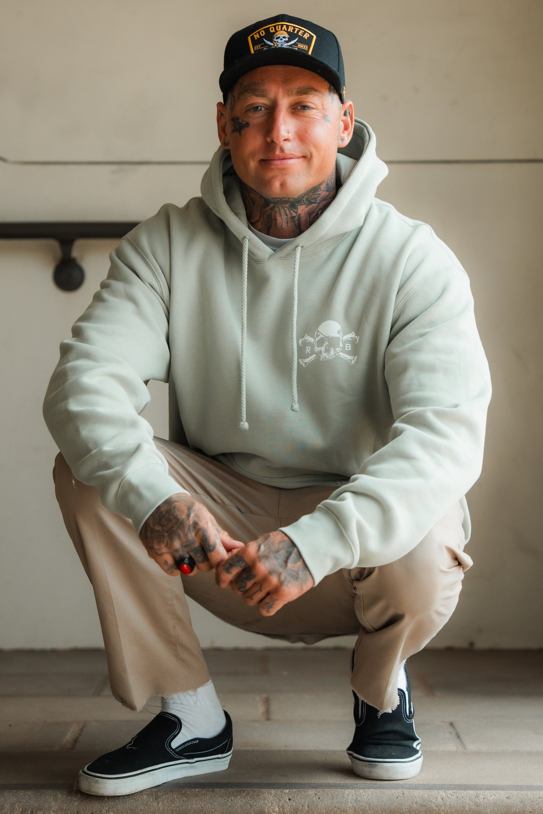 A man with tattoos squats on the ground, wearing a Relentless Betrayal Tell No Tales Premium Sage Hoodie, beige pants, black slip-on shoes, white socks, and a black cap. He smiles slightly at the camera.