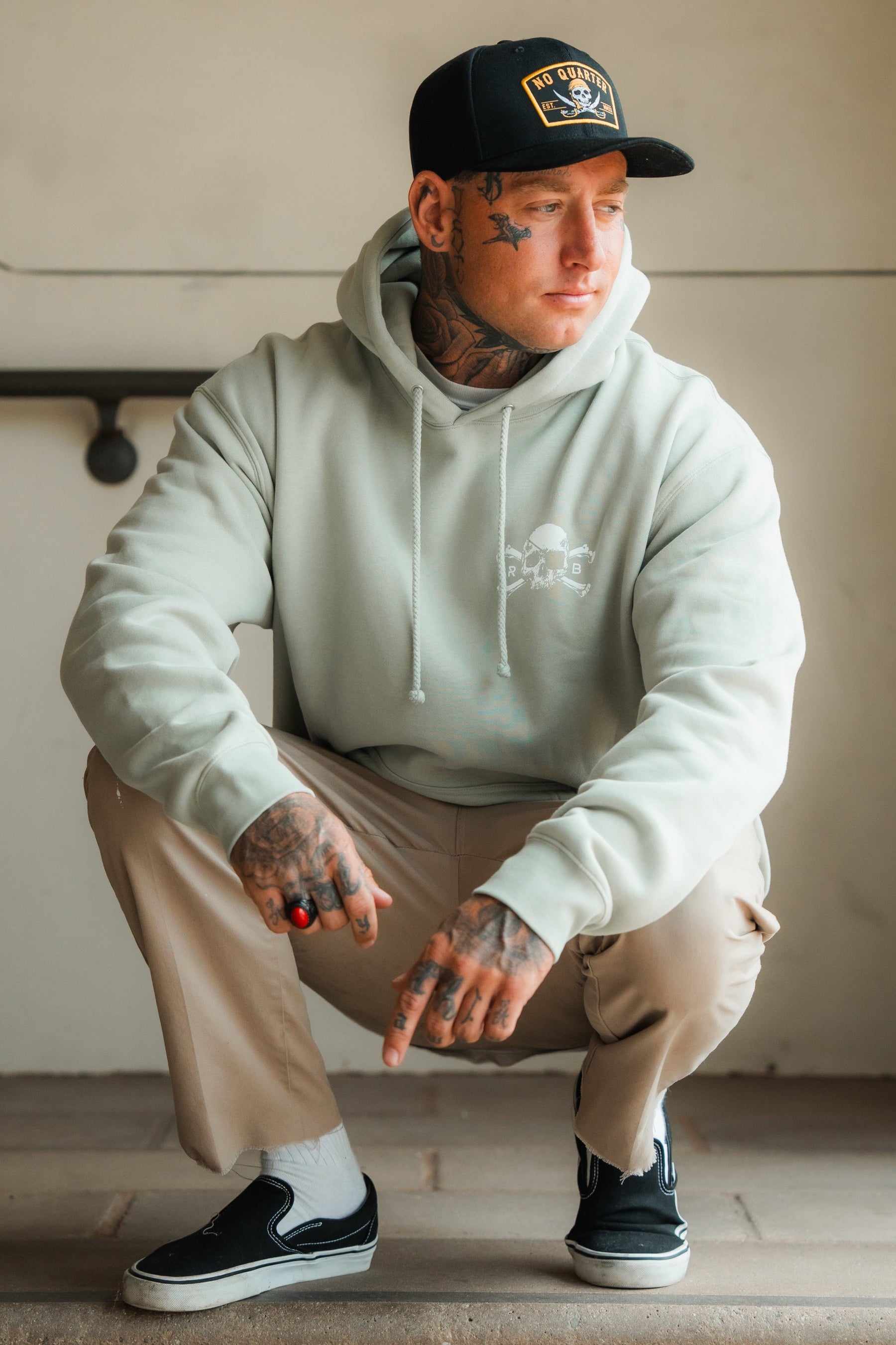A tattooed man squats indoors wearing the Relentless Betrayal Tell No Tales Premium Sage Hoodie, beige pants, a black cap, and black slip-on shoes. He looks to the side, with one hand on his knee and the other pointing down.