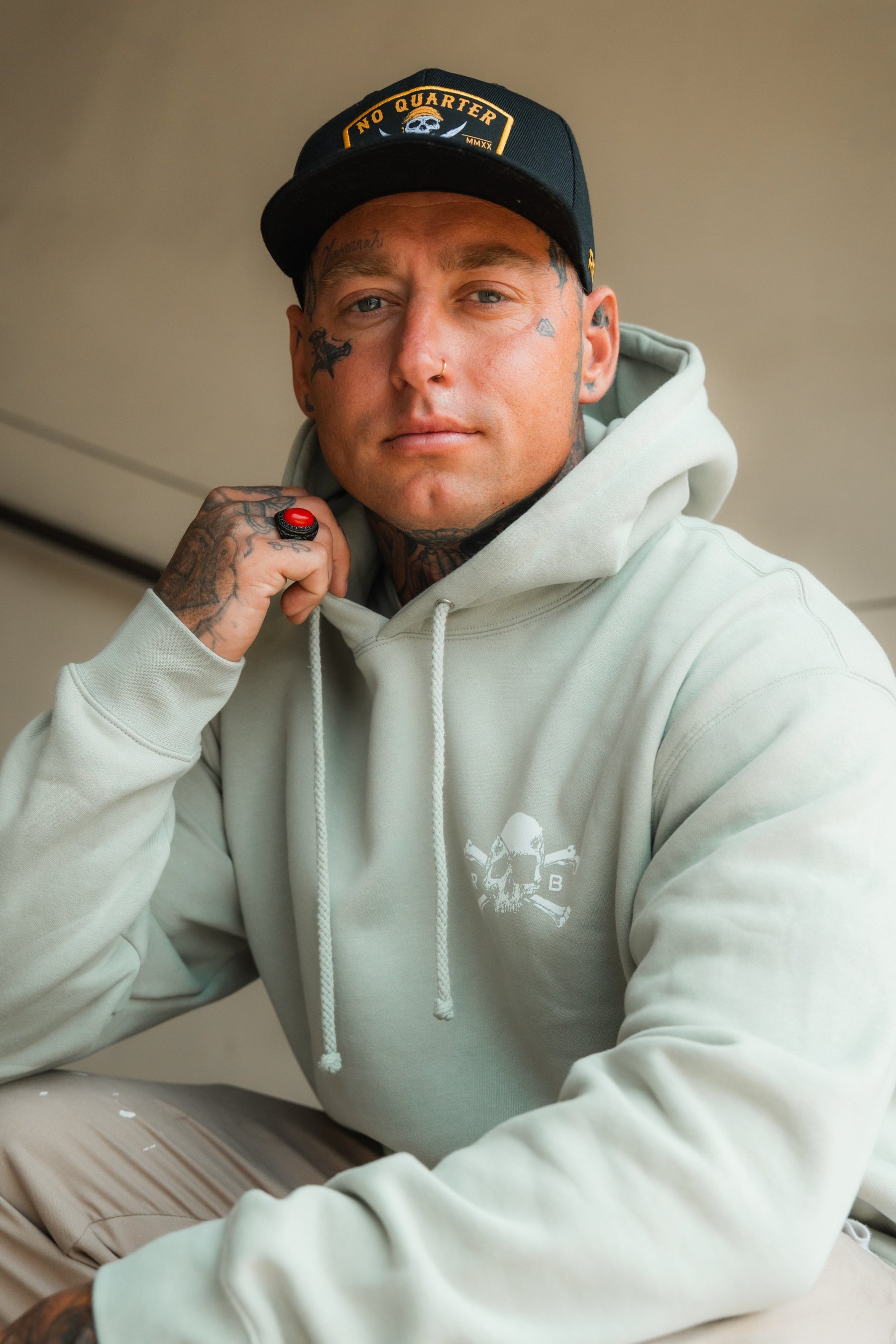 A man with face and hand tattoos wears a black No Quarter cap, beige pants, and the Relentless Betrayal Tell No Tales Premium Sage Hoodie, resting his ringed hand on his chin.