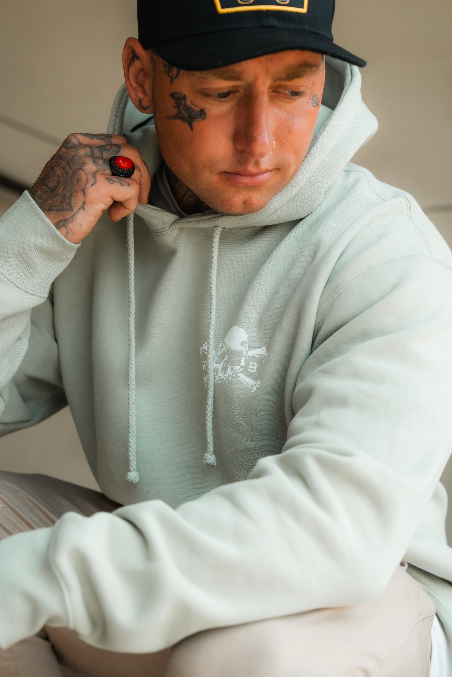 Wearing the Relentless Betrayal Tell No Tales Premium Sage Hoodie, a person with visible tattoos and a red ring looks down thoughtfully, hand near their neck. A black cap completes this unisex streetwear vibe.