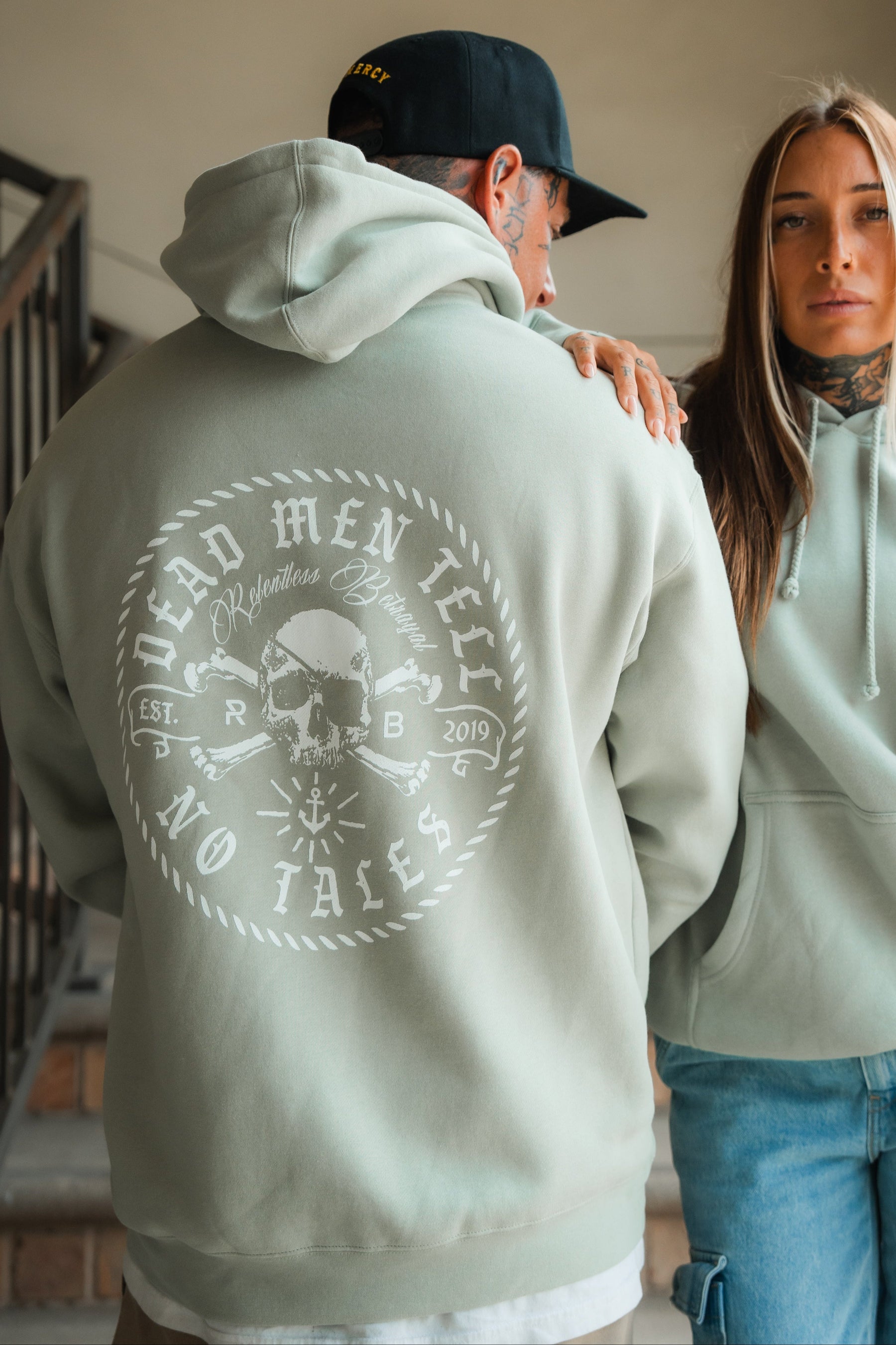 A man in a Relentless Betrayal Tell No Tales Premium Sage Hoodie stands next to a woman in a matching cotton-polyester blend hoodie. Both display tattoos and relaxed expressions as they pose indoors near stairs.