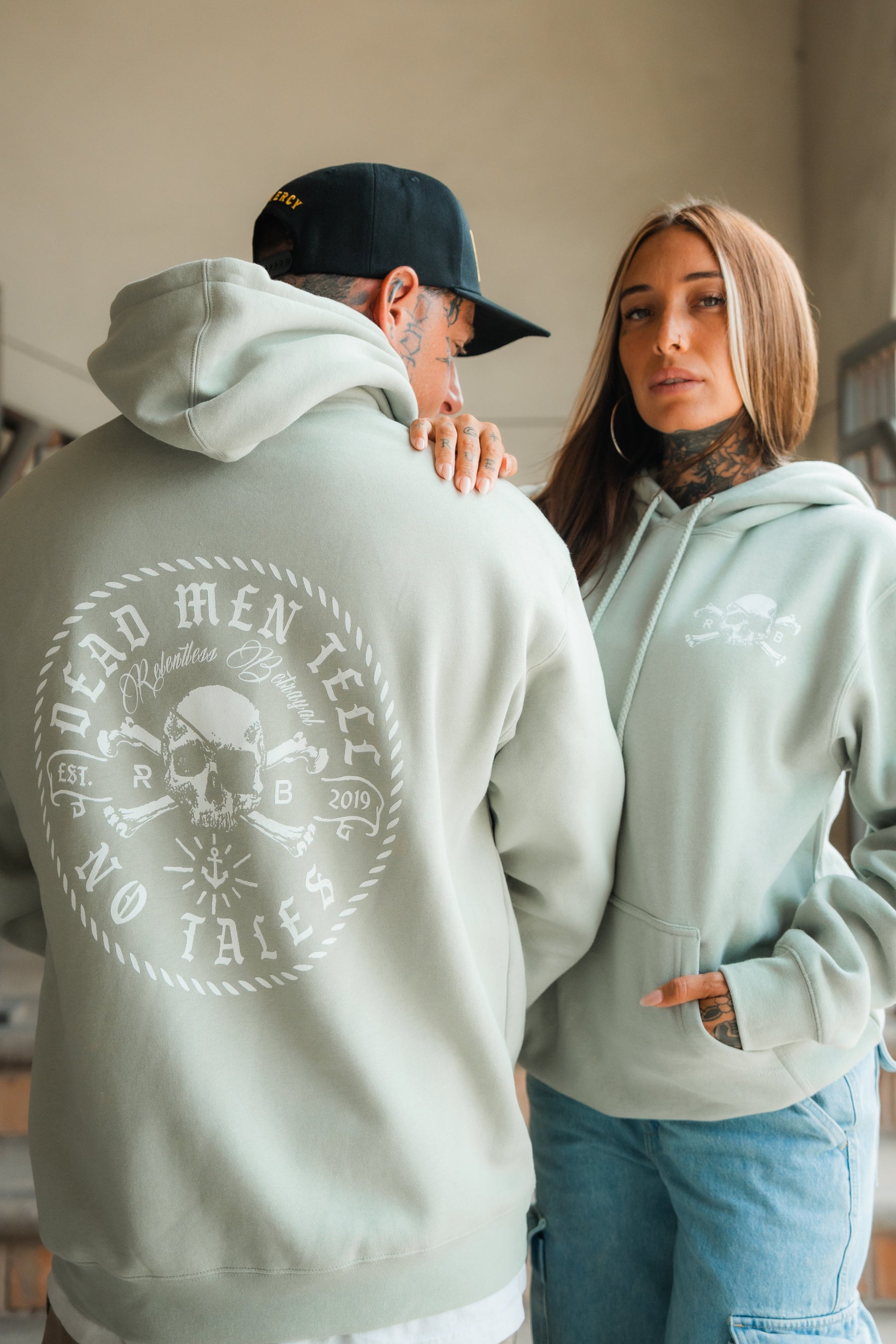 Two people wear Relentless Betrayal Tell No Tales Premium Sage Hoodies. The man faces away, showing the bold back graphic; the woman faces forward, her hand on his shoulder, revealing the subtle front logo.