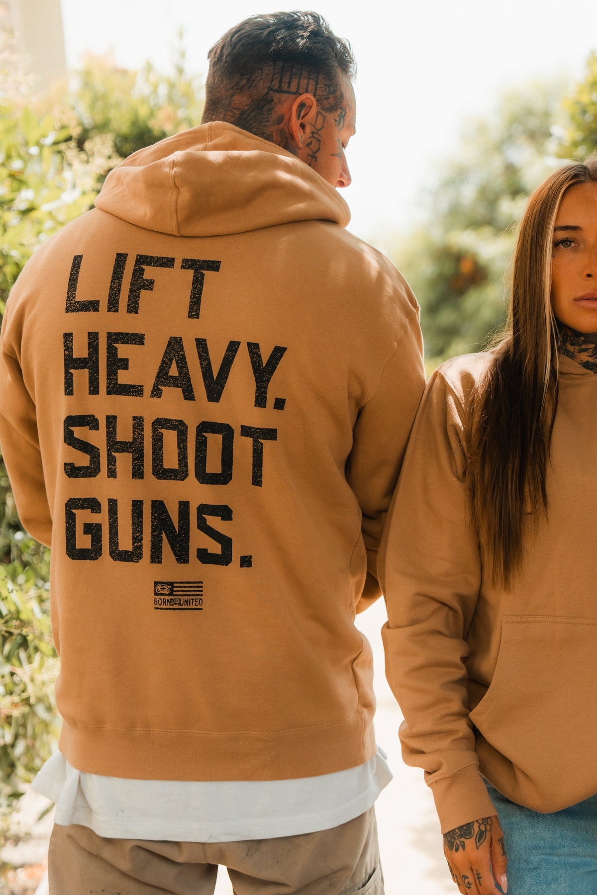 A man and woman in tan hoodies stand outdoors, both with tattoos and neutral expressions. The mans hoodie is the LHSG - Hoodie by Tango Charlie Apparel, featuring the bold Lift Heavy Shoot Guns motto—perfect for fitness and 2A enthusiasts.