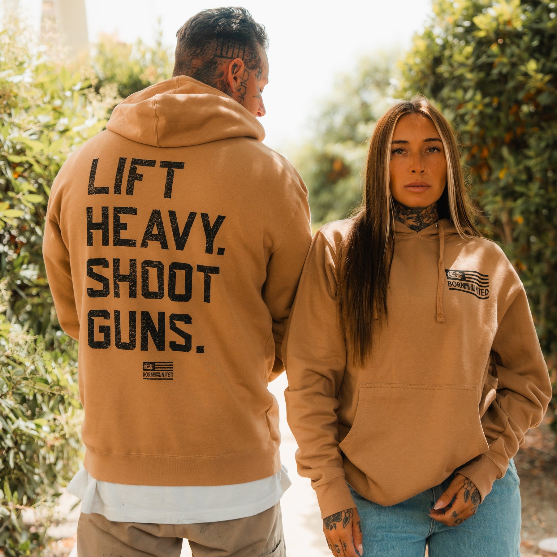 A man and woman in tan hoodies stand outdoors, both with tattoos and neutral expressions. The mans hoodie is the LHSG - Hoodie by Tango Charlie Apparel, featuring the bold Lift Heavy Shoot Guns motto—perfect for fitness and 2A enthusiasts.