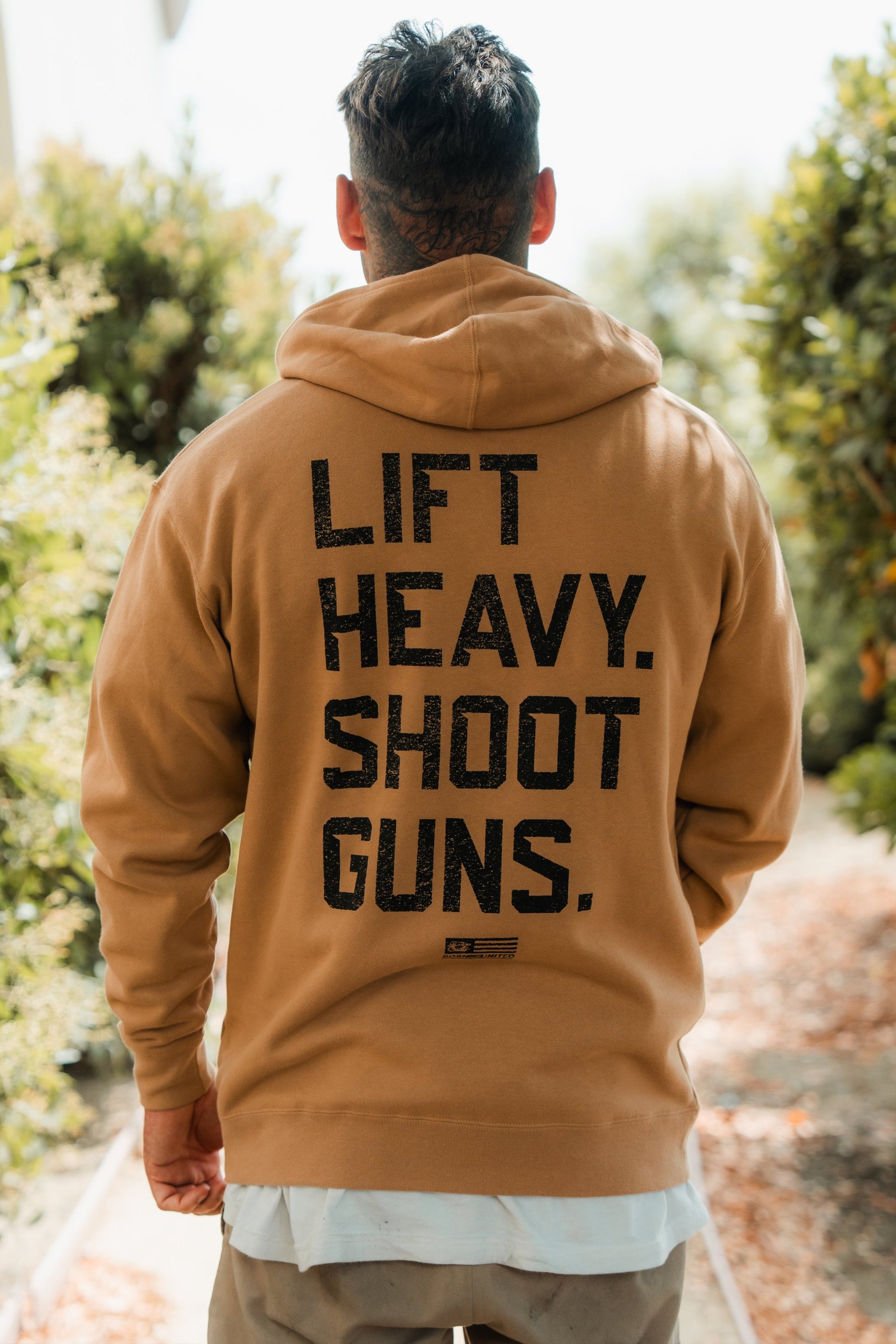 A person outdoors, back to the camera, wears the LHSG - Hoodie by Tango Charlie Apparel. The tan hoodie features Lift Heavy Shoot Guns, echoing bold, Second Amendment-inspired fitness style. Green bushes and a pathway complete the scene.