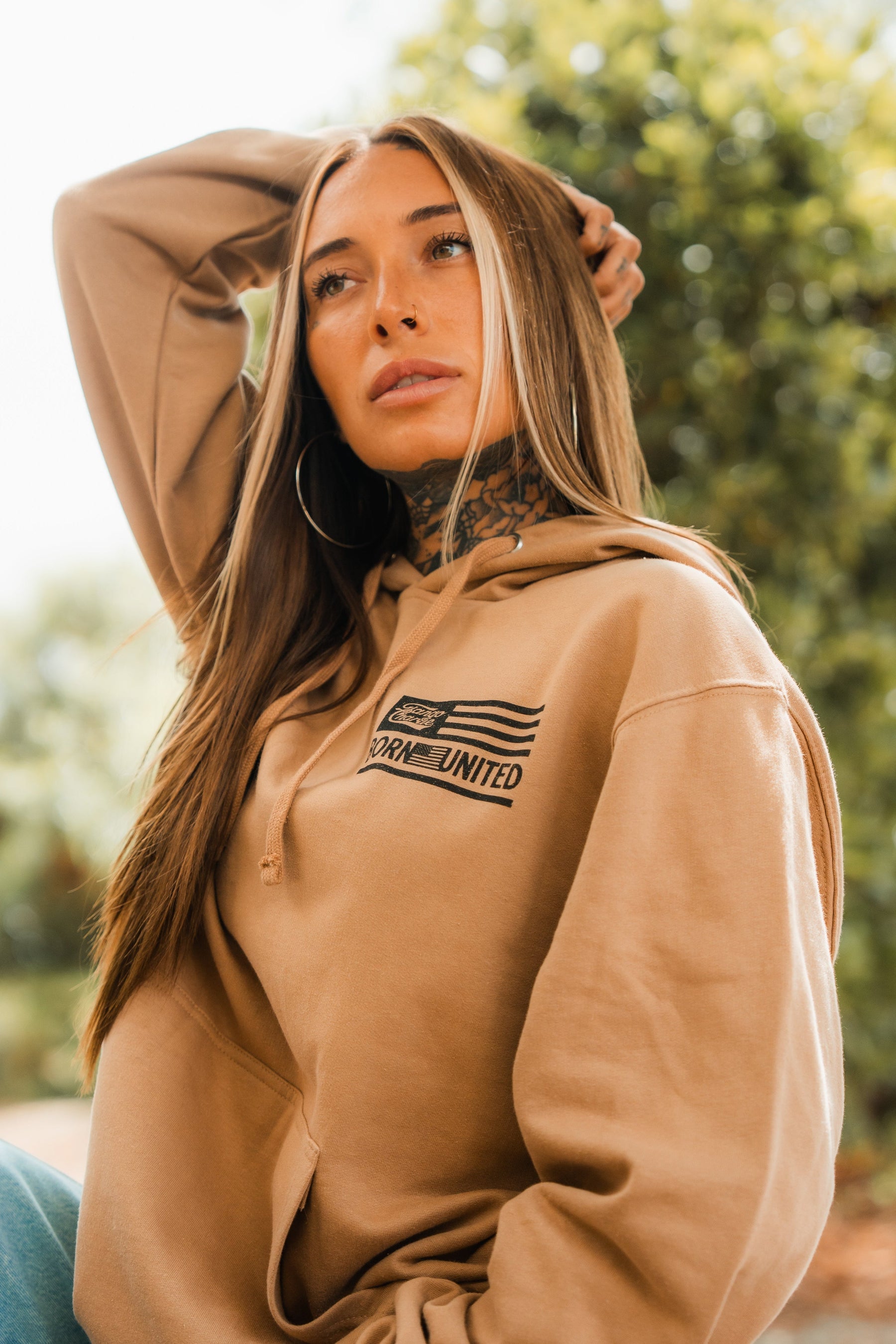 A woman with long straight hair and neck tattoos poses outdoors in Tango Charlie Apparel’s tan LHSG - Hoodie, branded with “Born United,” standing against a blurred green background.