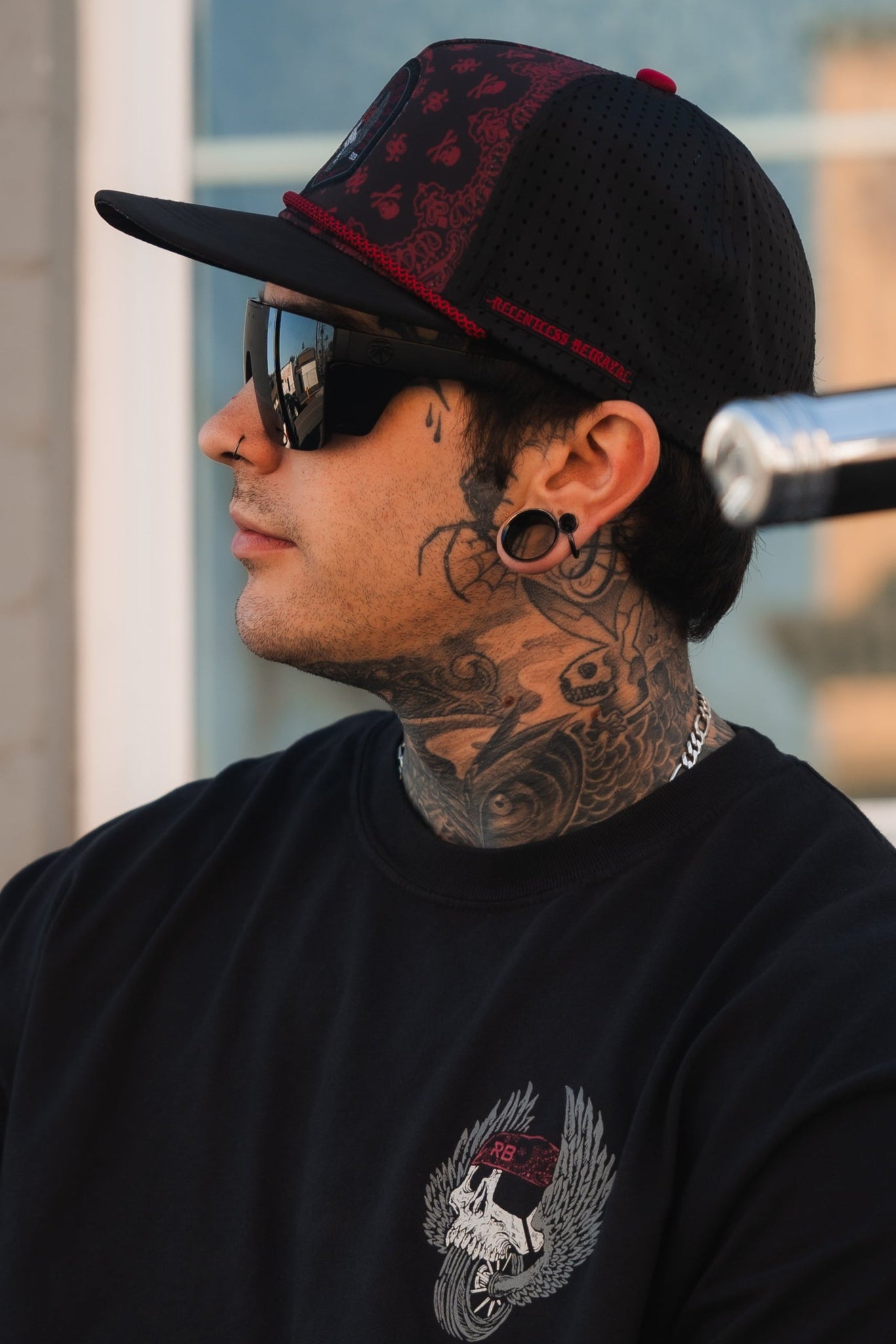 A person wearing the All Gas No Brakes Premium Snapback in black by Relentless Betrayal and large sunglasses is shown in profile. They have multiple facial piercings, extensive neck tattoos, and a black shirt with a winged emblem.