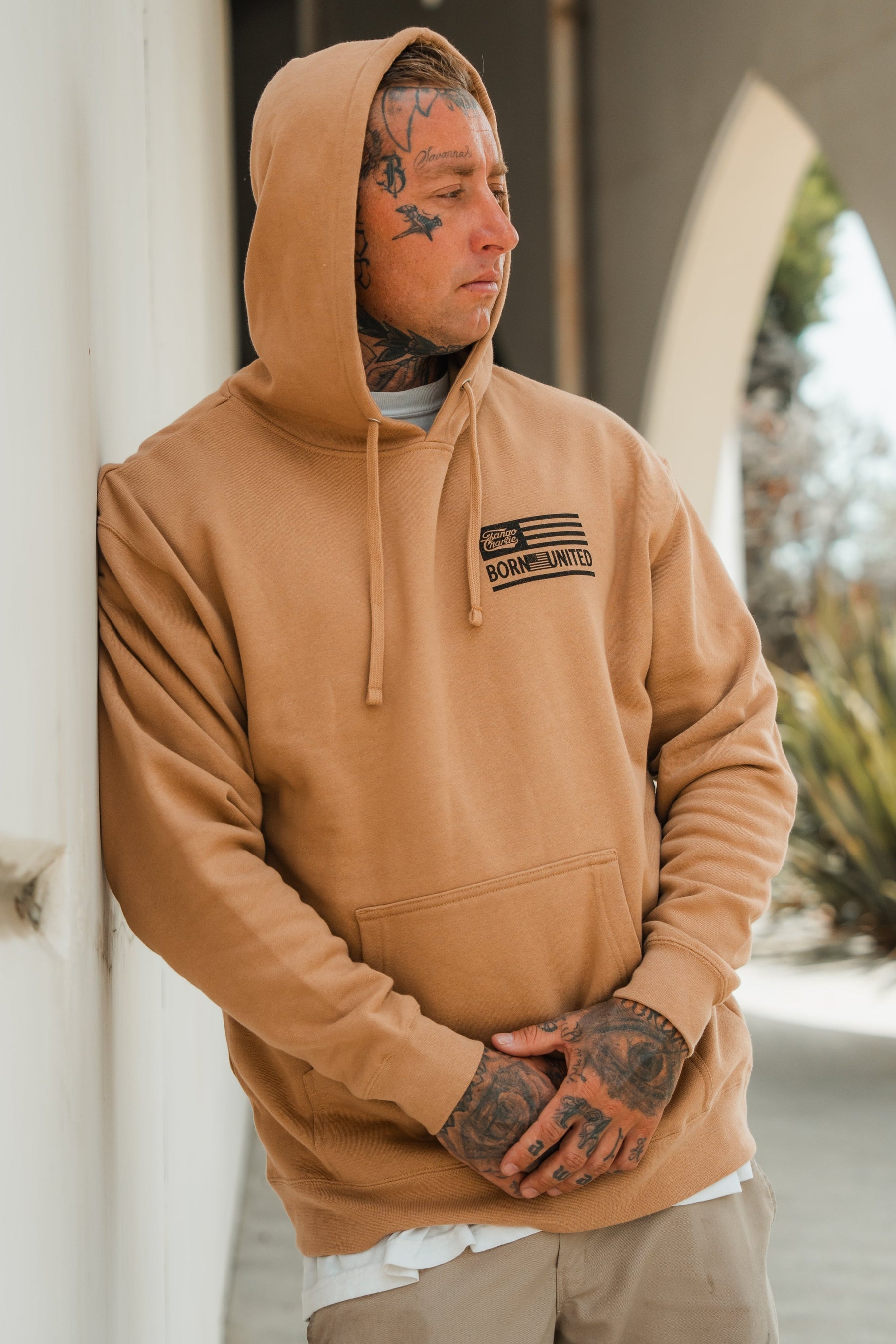 A tattooed person in a tan LHSG - Hoodie by Tango Charlie Apparel, featuring a small American flag, Born United text, and Lift Heavy Shoot Guns slogan, stands outdoors leaning against a wall and looking to the side.