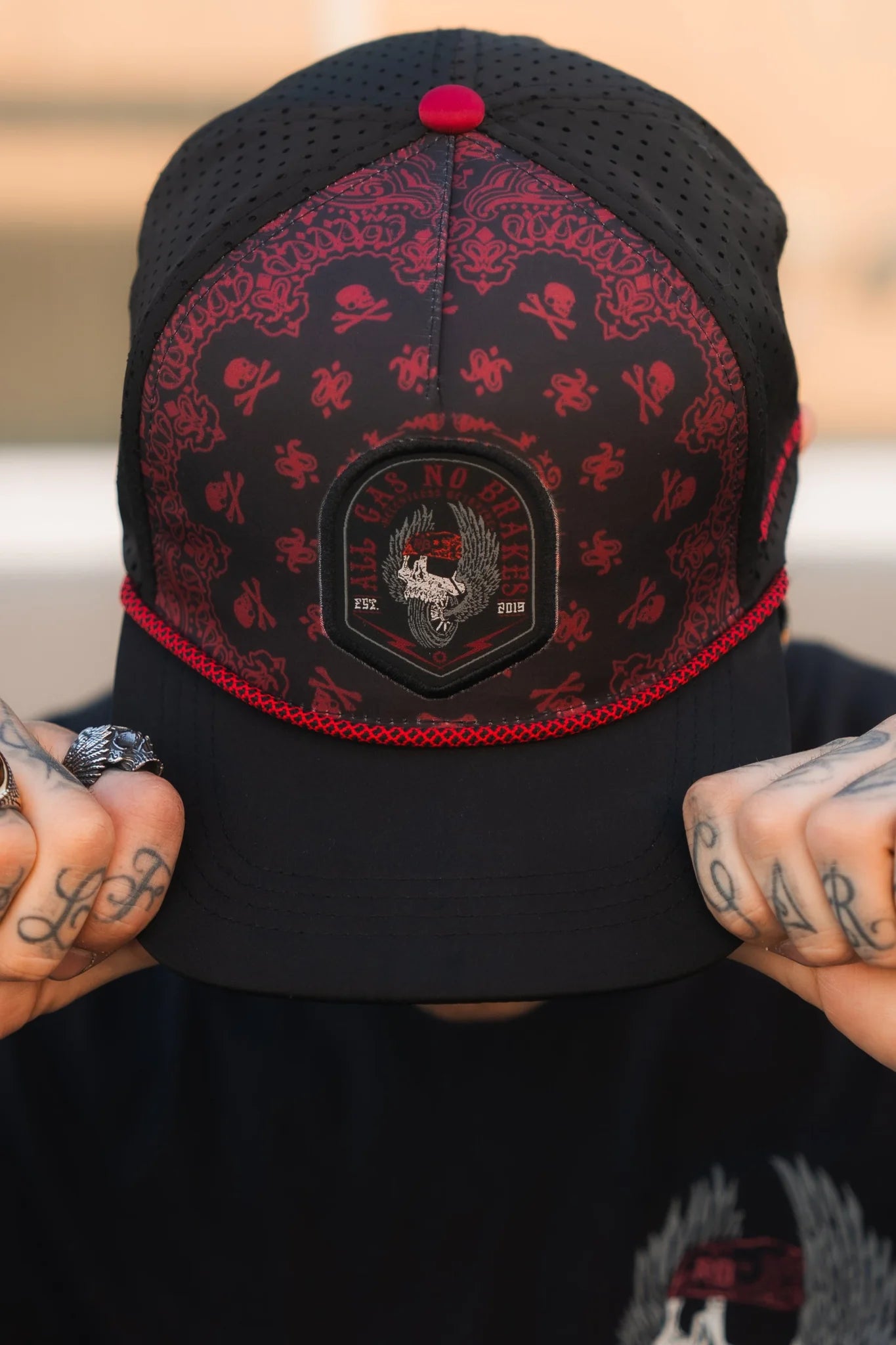 A person wearing the All Gas No Brakes Premium Snapback by Relentless Betrayal, featuring a black and red skull-and-crossbones design and tattooed fingers on the brim, sports a winged skull with text on the front. Bandana print accents add flair as the background fades into a blur.
