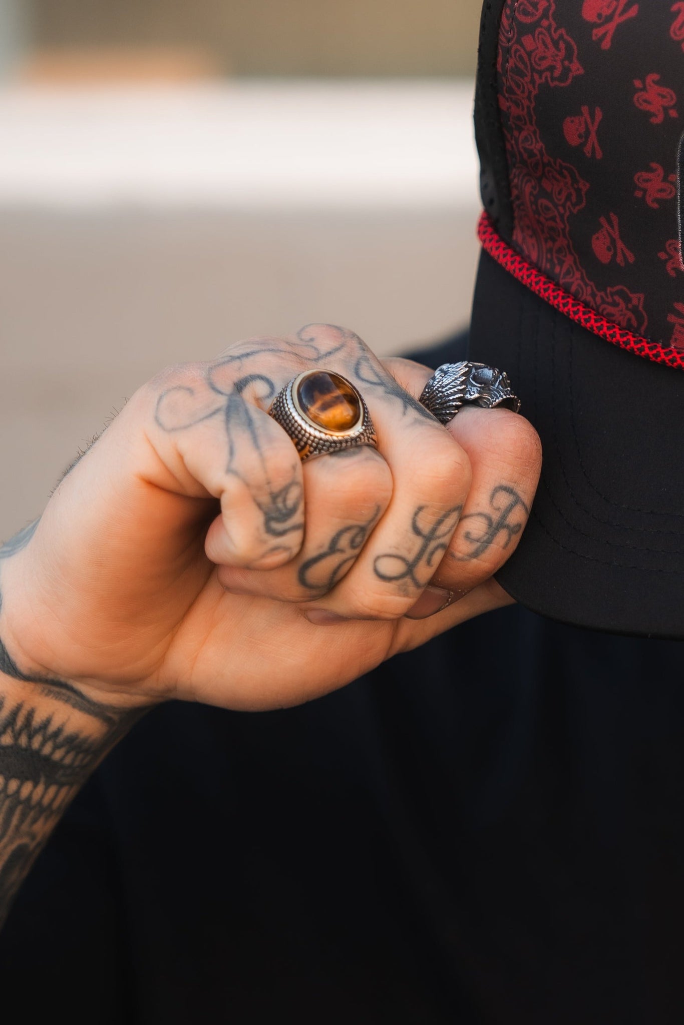 A tattooed hand, adorned with rings including a handcrafted stainless steel one featuring tigers eye and the Relentless Betrayal Inferno Ring with a striking wolf head, grips the brim of a black cap detailed with red stitching and patterns.