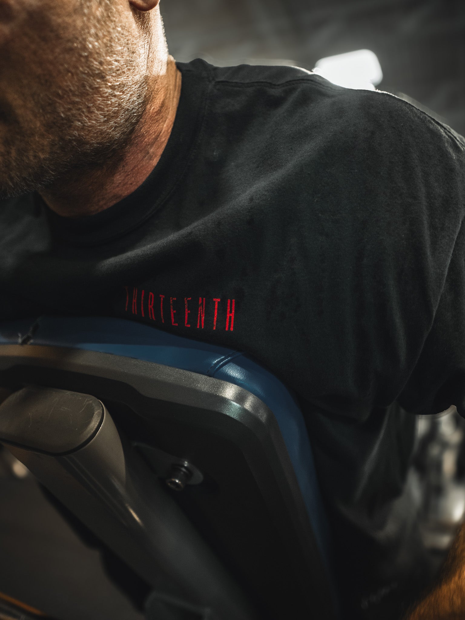 A man wearing the black 13th Samson Premium Tee with THIRTEENTH in red exercises on gym equipment, with the image highlighting his upper torso and part of his face.