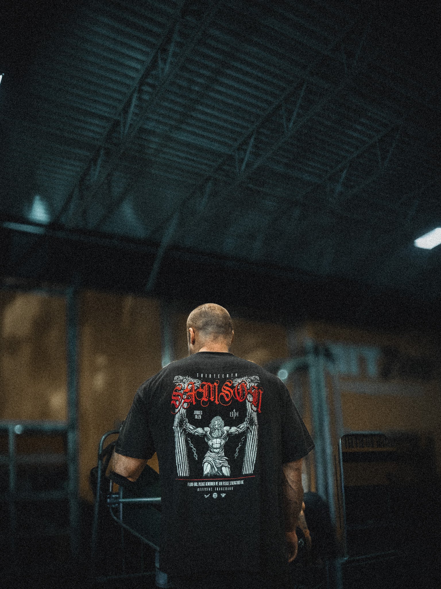 A person with a shaved head stands indoors, facing away, wearing the 13th Samson Premium Tee featuring a bold graphic and red text on the back. The dim lighting and industrial setting accentuate this premium garment.