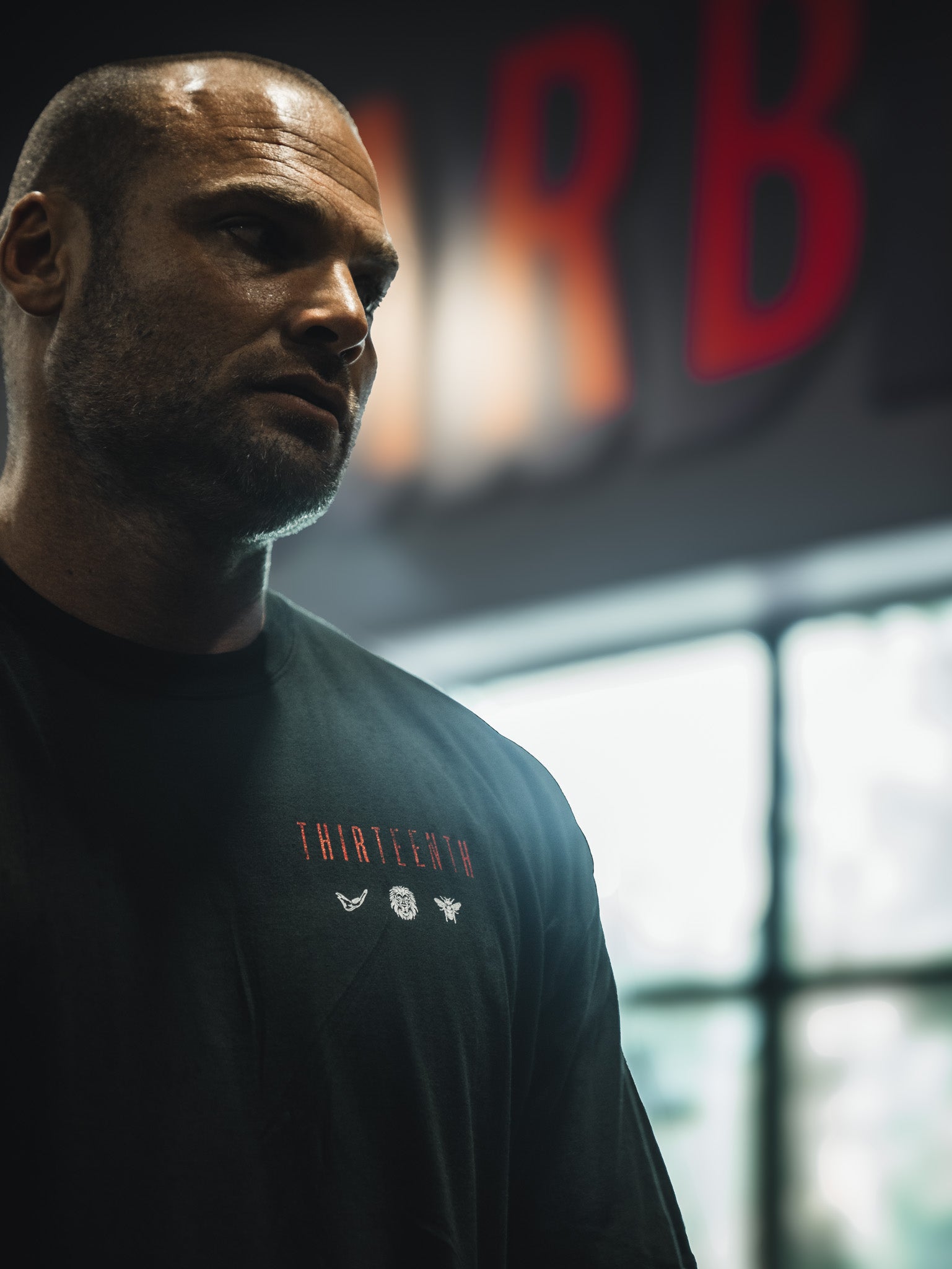 A man with a shaved head stands indoors before a blurred window, wearing the 13th Samson Premium Tee featuring THIRTEENTH and small graphics. The dim background shows red, out-of-focus letters.