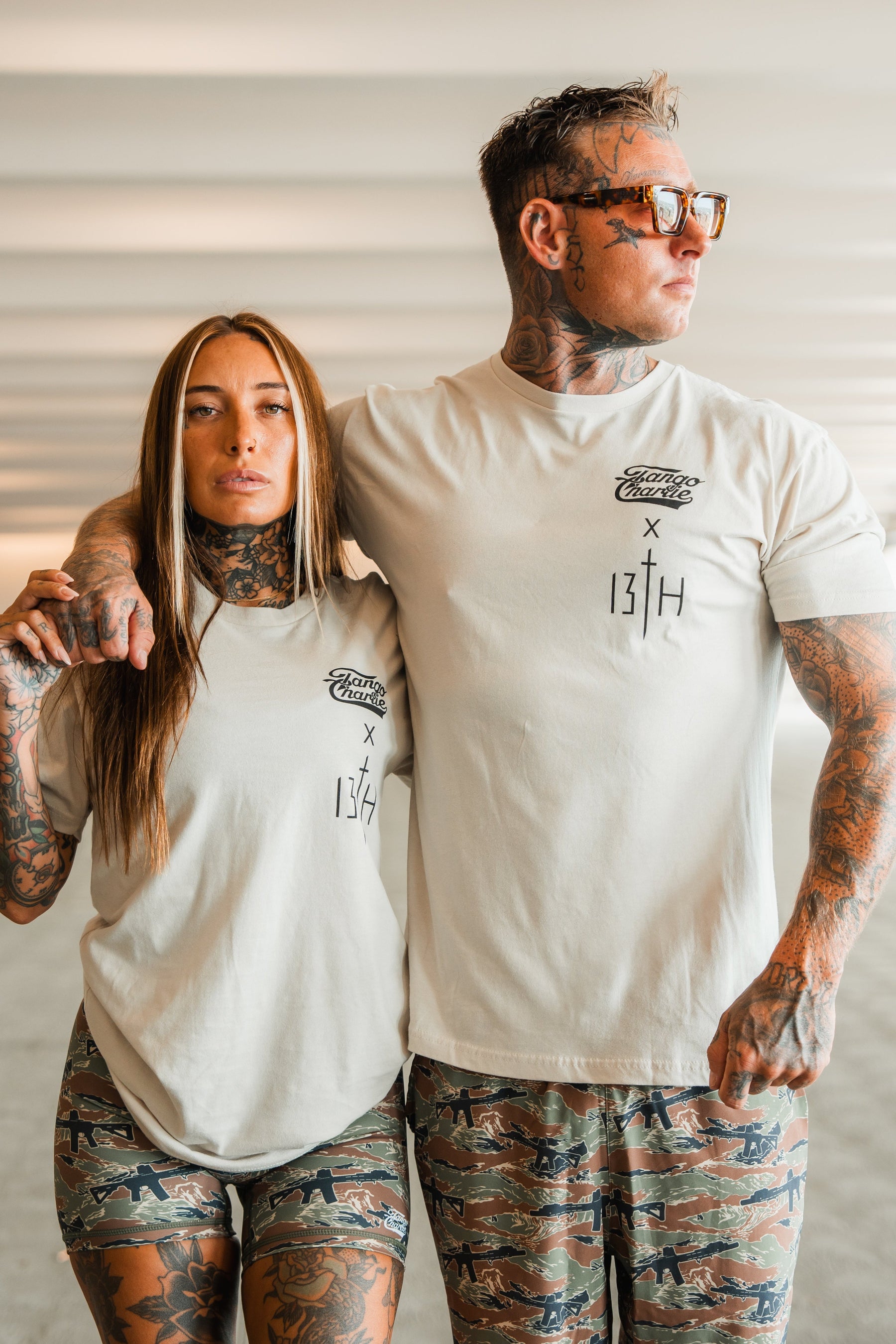 A tattooed man and woman wear matching Lift Heavy. Pray Hard. tees from Tango Charlie Apparel, standing side by side in beige shirts and camo shorts, exuding spiritual resilience and strength together.