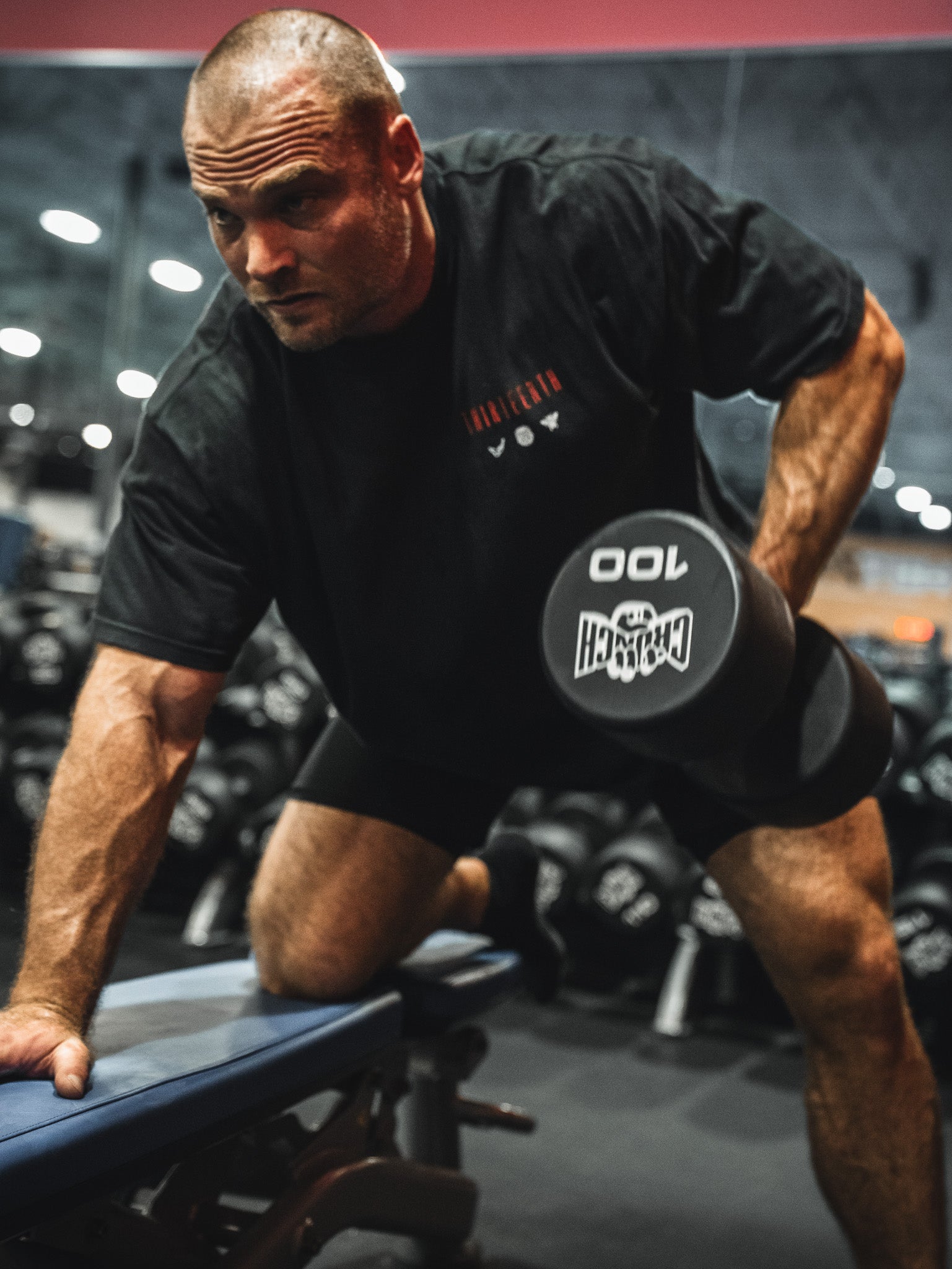 A muscular man in a black 13th Samson Premium Tee and shorts performs a one-arm dumbbell row with a 100-pound weight on a gym bench, surrounded by mirrors and racks of dumbbells.